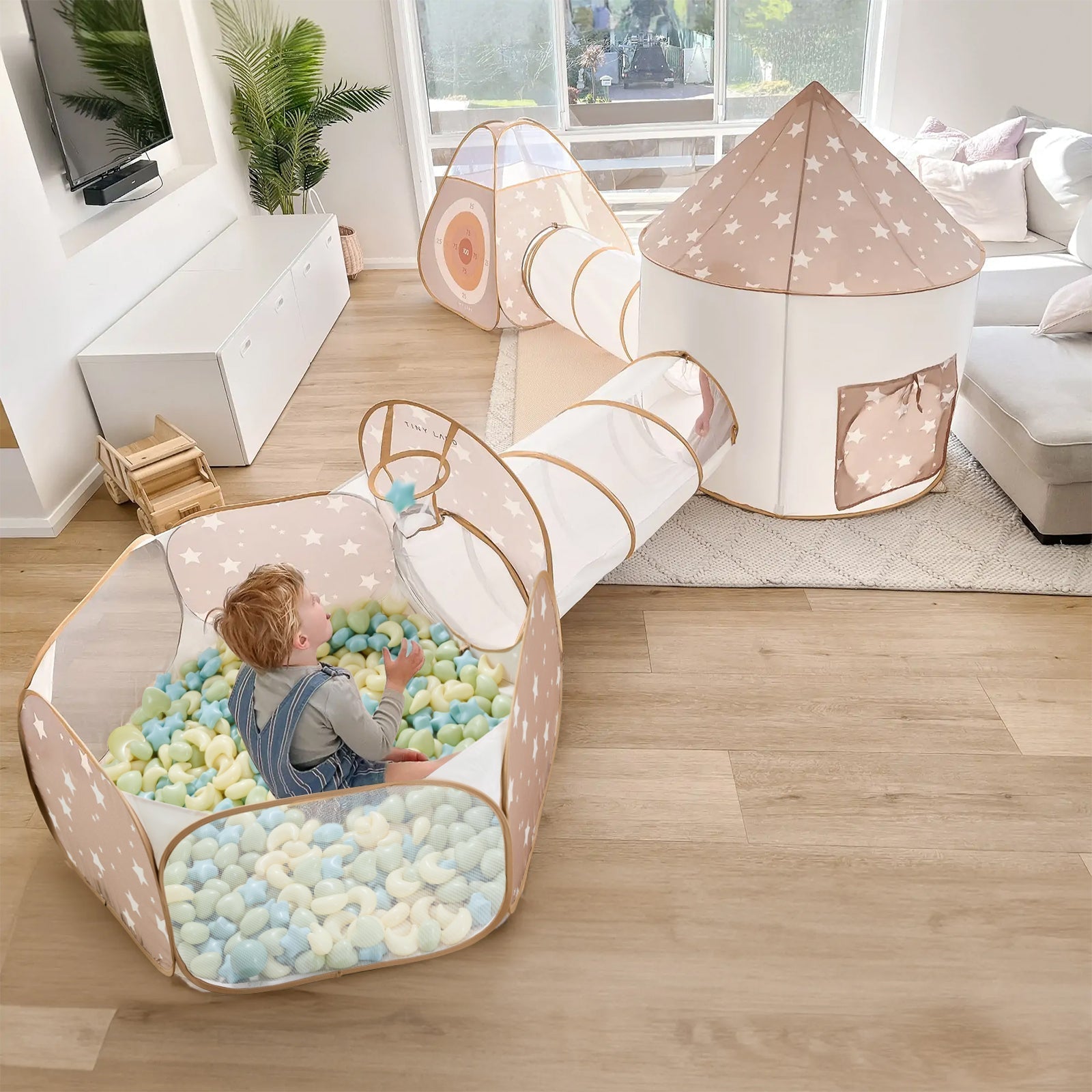 A little boy sits in a bright living room, playing with an extra-large crawl tunnel, ball pit, and ball pit balls.