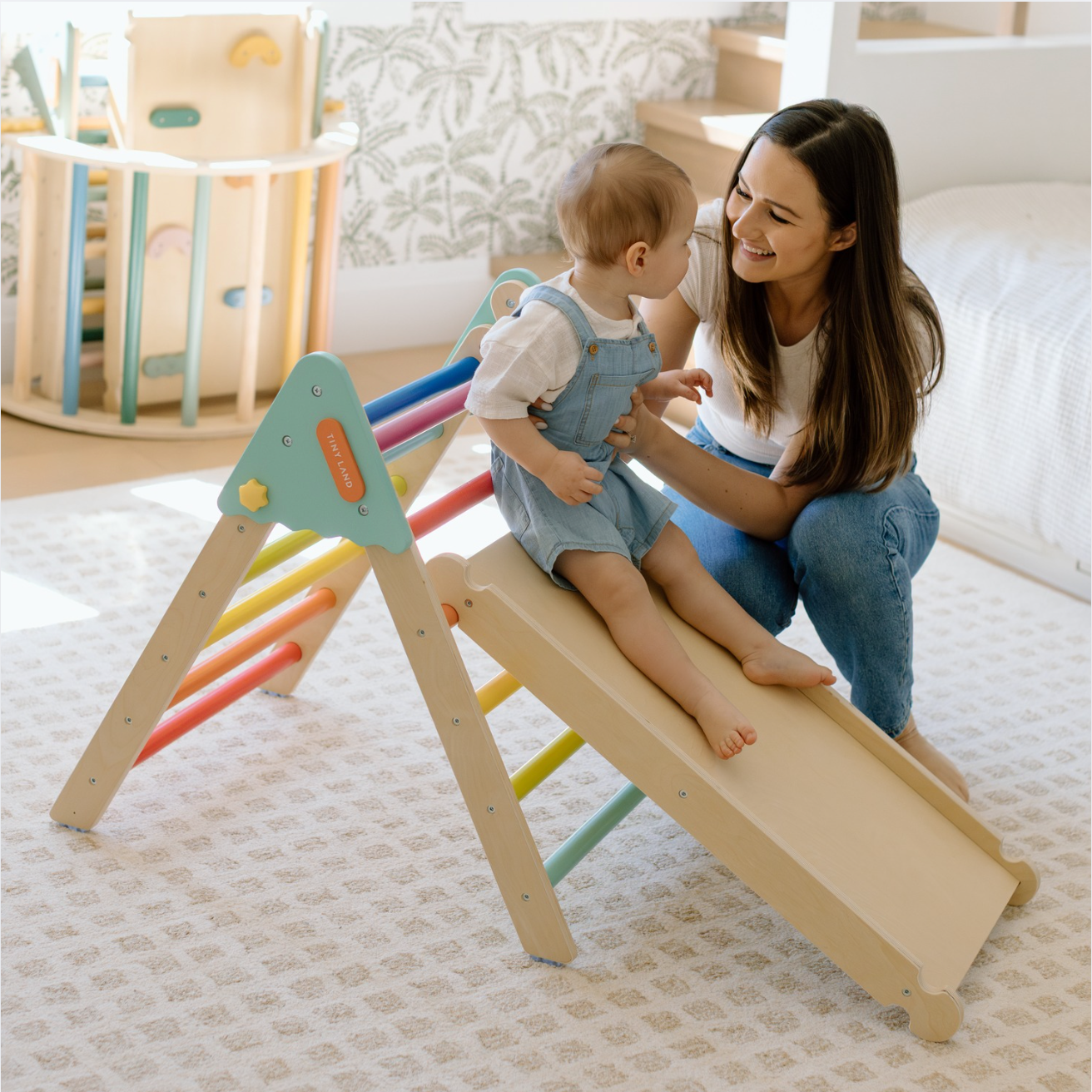 In the playroom, a black-haired mom in a short-sleeve top and long pants happily plays with her little one on the colorful climbing frame.