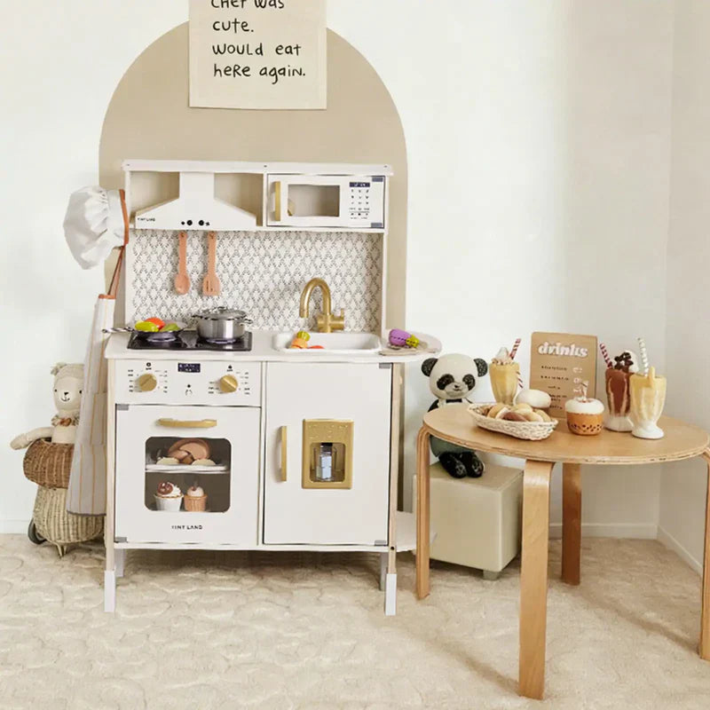 A white wooden toy kitchen is placed in the playroom, filled with toy food, kitchen utensils, a chef’s apron, and a hat.