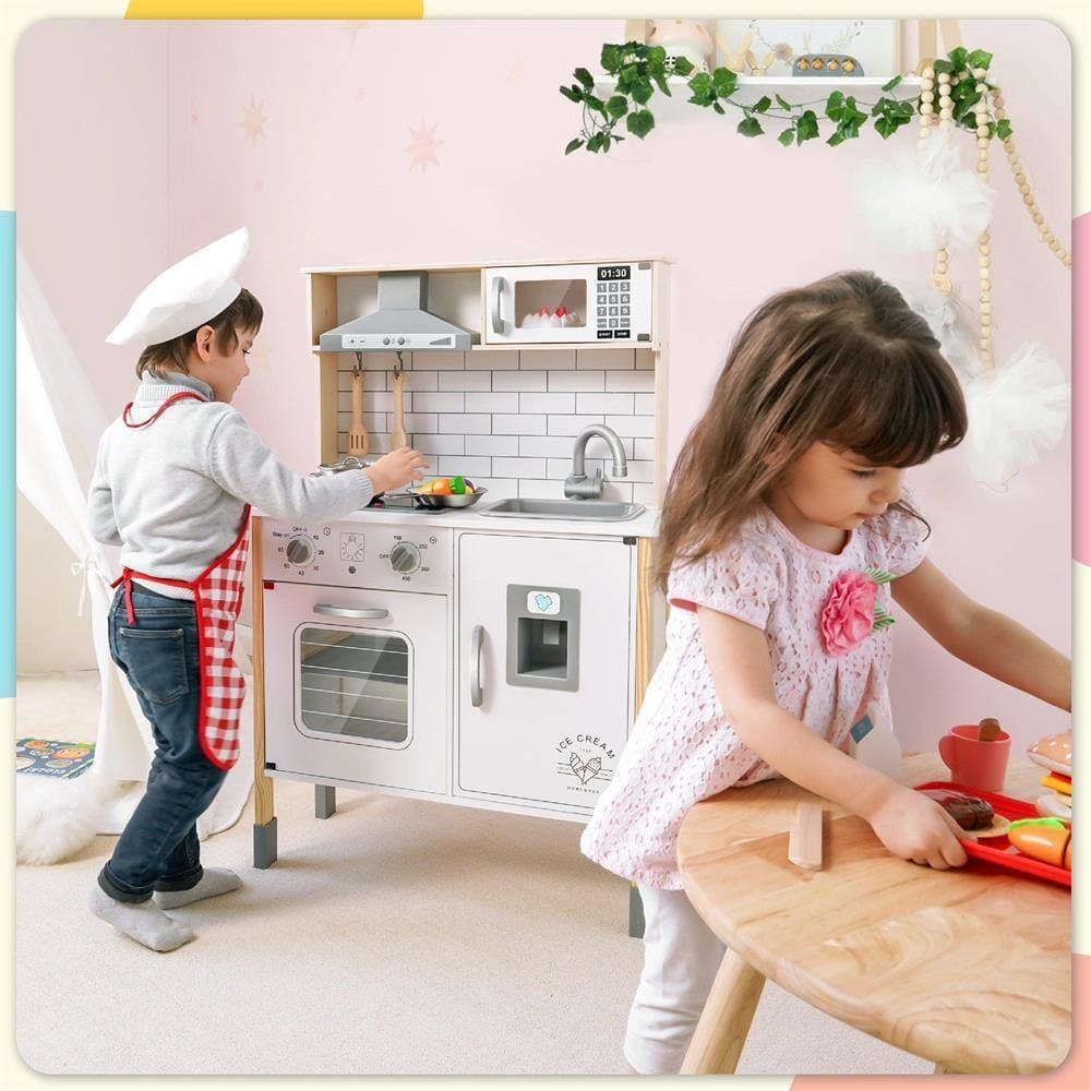 A little boy wearing an apron and chef’s hat is cooking in the toy kitchen, while a little girl in a dress is playing with play food at the nearby table.