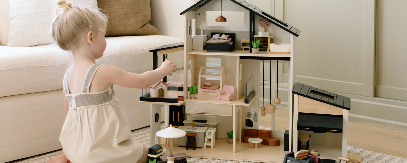 A young child in a light dress sits on the floor, playing with a wooden dollhouse filled with miniature furniture in a cozy, softly lit living room.