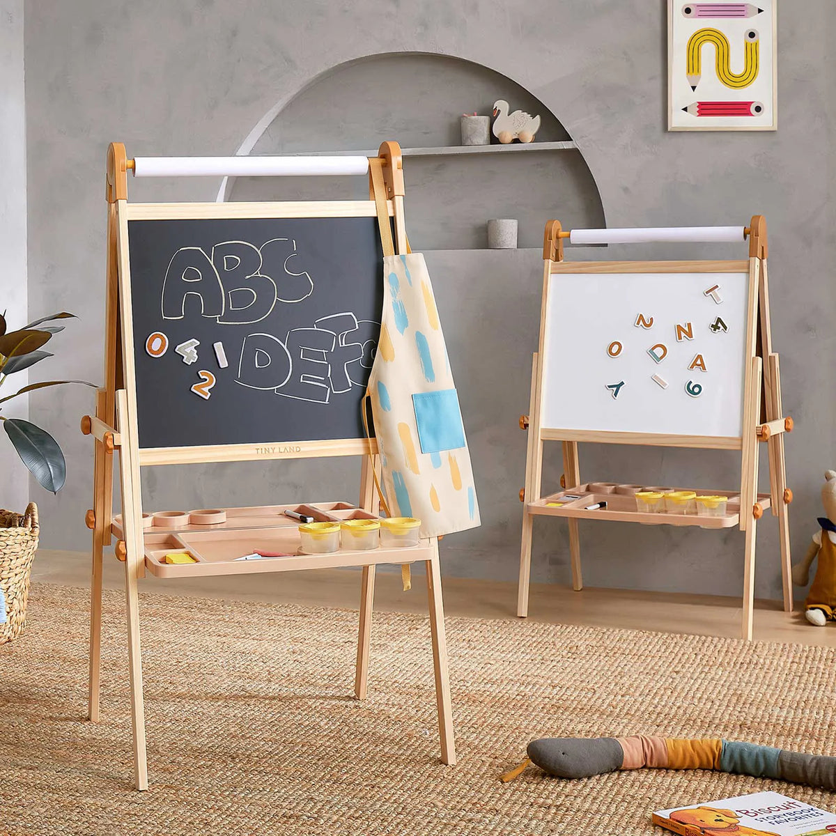 Two Tiny Land® Caramel Magnitales Easels for Kids by Tiny Land stand in a cozy playroom. One displays chalk letters on its chalkboard side, while the other serves as a magnetic easel with colorful magnetic letters. Art supplies and a patterned apron hang from the adjustable easels, surrounded by soft toys and cheerful decor throughout the room.