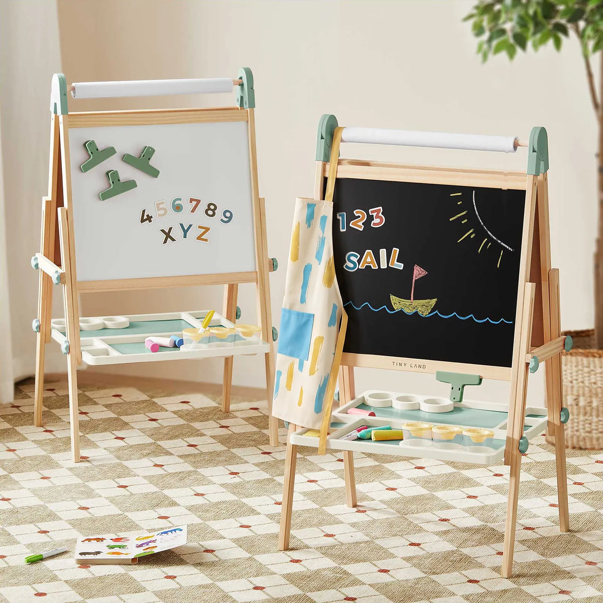 Two Tiny Land® Magnitales Easels for Kids by Tiny Land, each equipped with a tray, display a magnetic whiteboard and a chalkboard featuring a sailboat drawing. Art supplies and a patterned cloth hang nearby, all arranged on top of a geometric rug.|Green