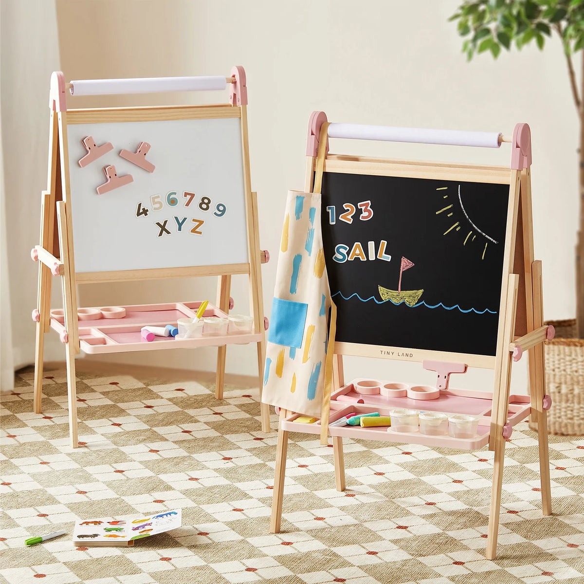 Two Tiny Land® Magnitales Easels for Kids by Tiny Land stand on a patterned rug. One magnetic easel showcases colorful letters and numbers, while the other adjustable art easel features a chalk drawing of a boat, numbers, and the word “SAIL.” Art supplies are neatly organized in trays below each board.|Pink