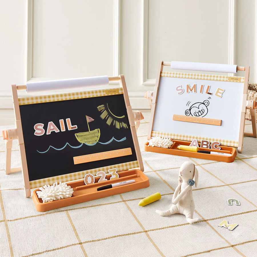Two Tiny LandĀ® Double-Sided Magnetic Tabletop Easels display colorful drawings and magnetic letters, setting up an inviting art station. A small plush bunny rests on the floor nearby, surrounded by chalk, erasers, and alphabet blocks in a cozy, sunlit room.|Oatmeal