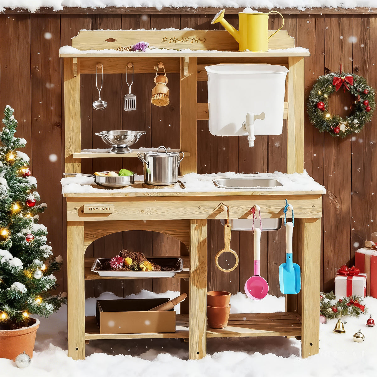 The Tiny Land® Mud Kitchen - DuoPlay Outdoor Play Kitchen for Kids by Tiny Land stands outdoors, topped with snow, adorned with utensils, a yellow watering can, and a wreath. Nearby are a small decorated Christmas tree and gifts for festive outdoor sensory play.