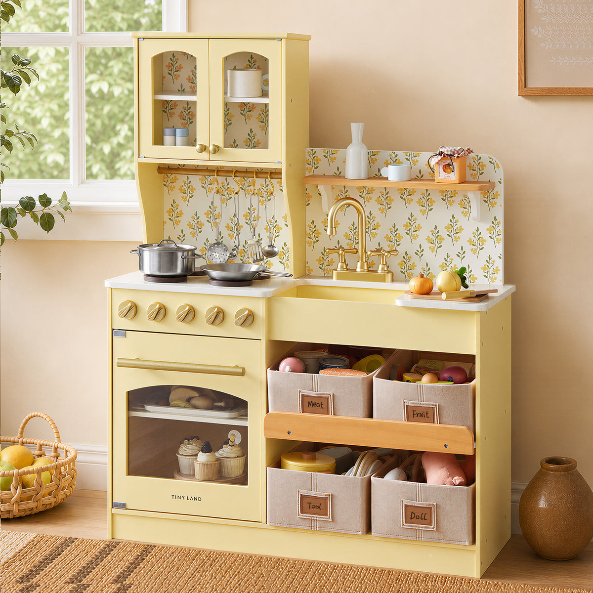 The Tiny Land® Wooden Play Kitchen with Storage for Creative Toddlers by Tiny Land, featuring multifunctional storage, play food, pretend cookware, and convenient baskets labeled Fruit, Vegetable, Tools, and Doll, sits against a wall adorned with a floral backsplash and a sunlit window—creating a cozy and eco-friendly play space.|Pale Yellow