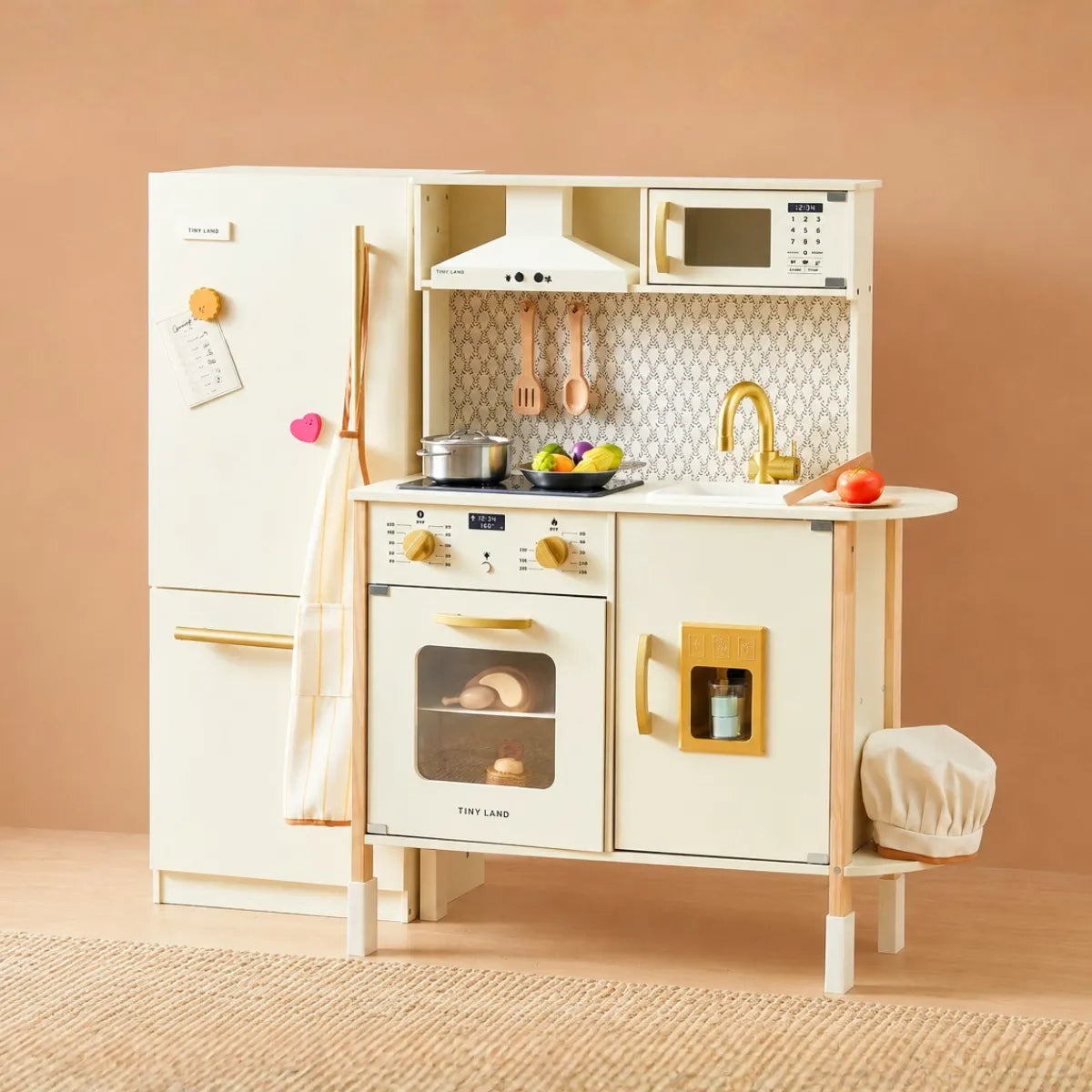 The Tiny Land® PopJoy Kitchen & Fridge Set by Tiny Land features a cream kids’ kitchen with fridge, oven, sink, microwave, and stove—plus toy food, utensils, and a towel—arranged on a woven rug for endless pretend play.