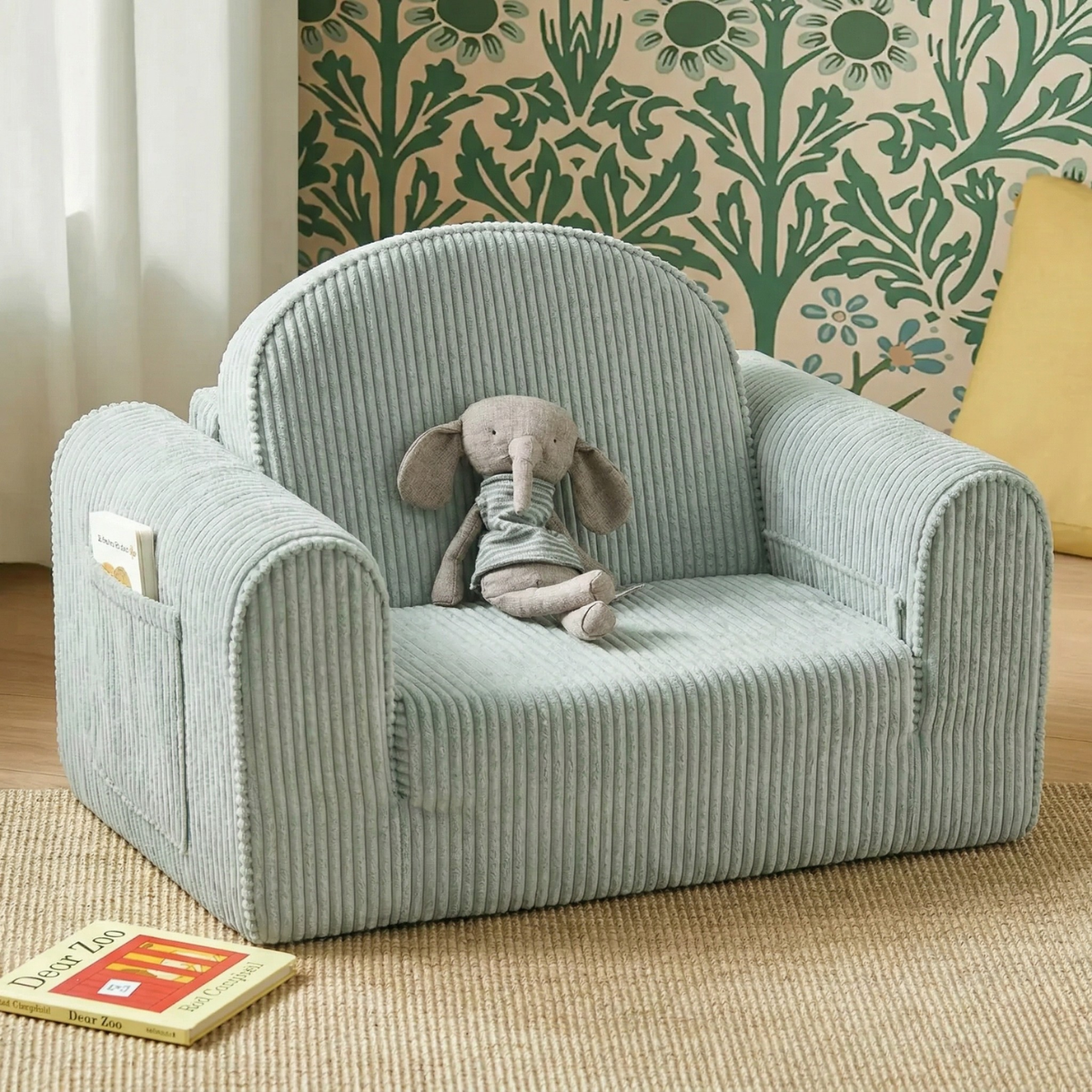 The Tiny Land® GentleSnug Kid Chair in blue is a cozy addition to any playroom, shown here with a plush elephant toy against green floral wallpaper. A yellow cushion and two children's books are placed nearby.