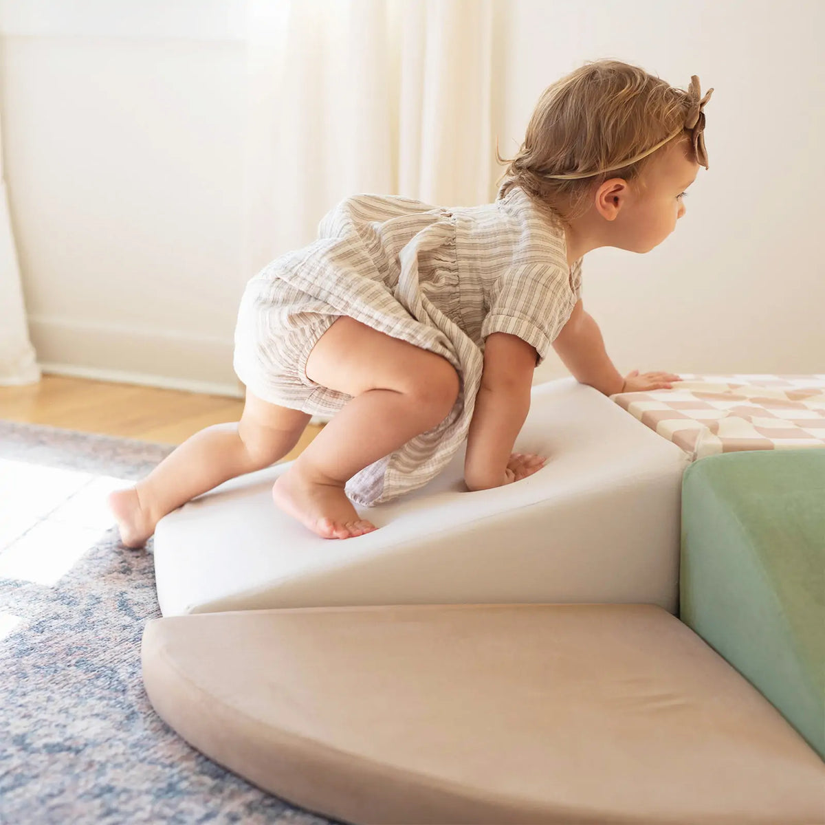 A toddler in a striped dress and headband climbs Tiny Land® PlayNest Climbing Blocks, boosting gross motor skills, in a sunlit room with light curtains and a rug.