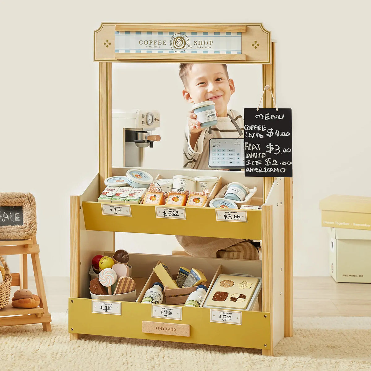 A smiling child stands behind the Tiny Land INC Farmers’ Market Gift Set, featuring a wooden market stall and organic food toys. Displaying play produce and prices, the cozy room is filled with cheerful pretend market fun.