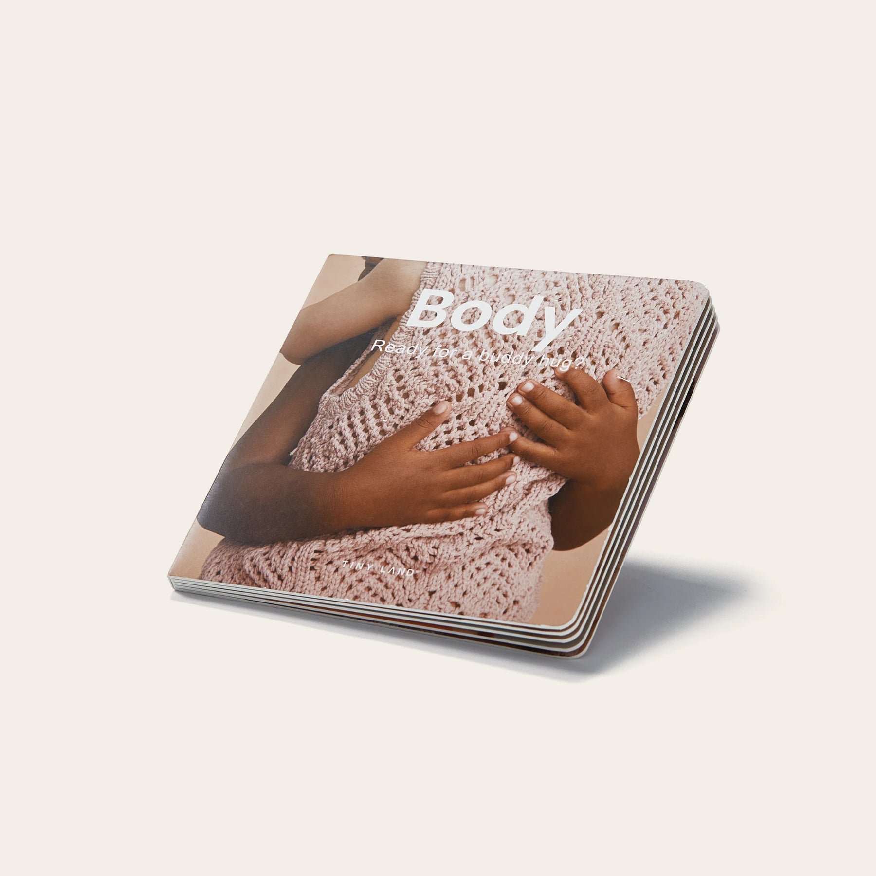 A small board book titled "Body" features a cover photo of two children’s hands touching a person in a light pink sweater, evoking the sensory exploration of Tiny Land® Montessori Toys for Newborns (7–9 Months) by Tiny Land. The background is plain and light-colored.