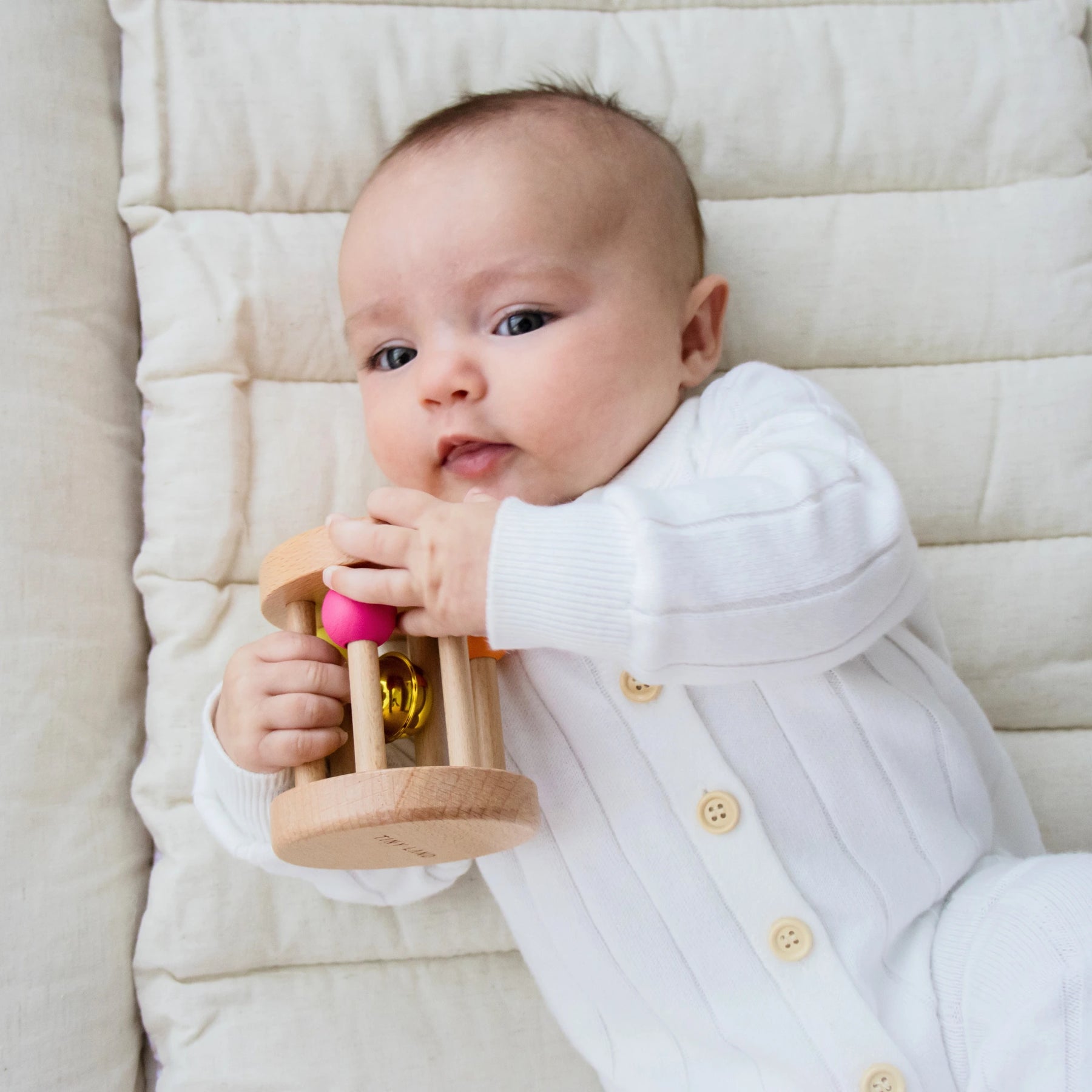 A baby in a white onesie lies on a padded cream-colored playmat, holding Tiny Land® Montessori Toys for Newborns (0–3 Months) from Tiny Land Official Store and looking up.