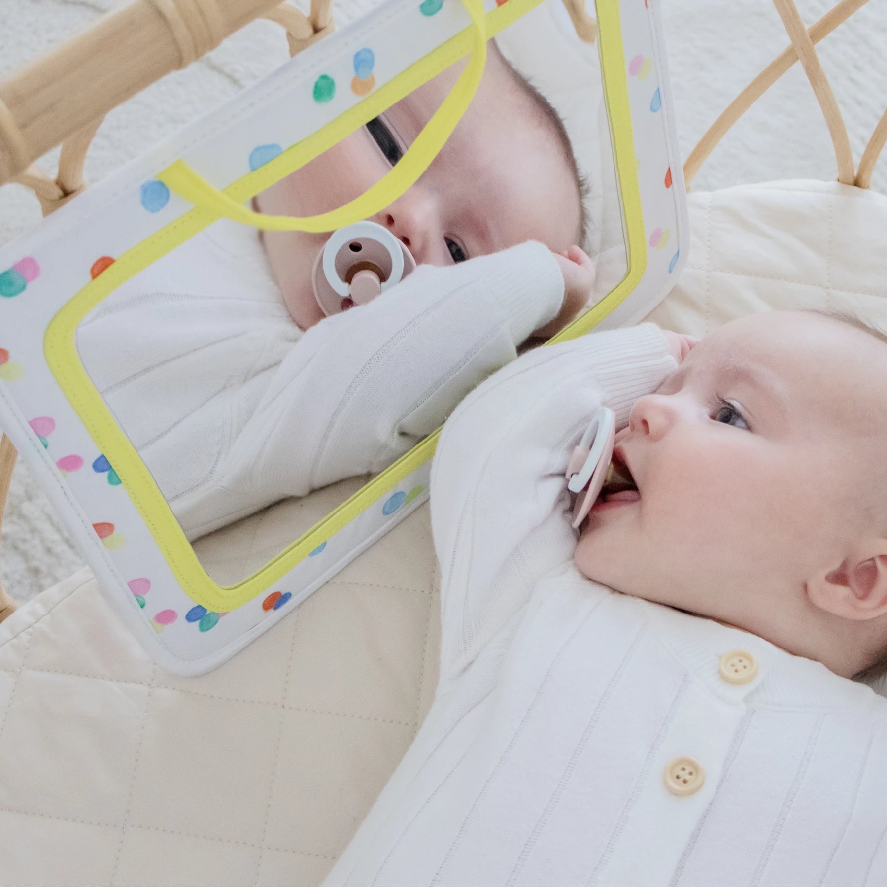 A baby in white lies on a quilted playmat, smiling at their reflection in a colorful mirror—a joyful moment with Tiny Land® Montessori Toys for Newborns (0–3 Months) from the Tiny Land Official Store, while holding a pacifier.