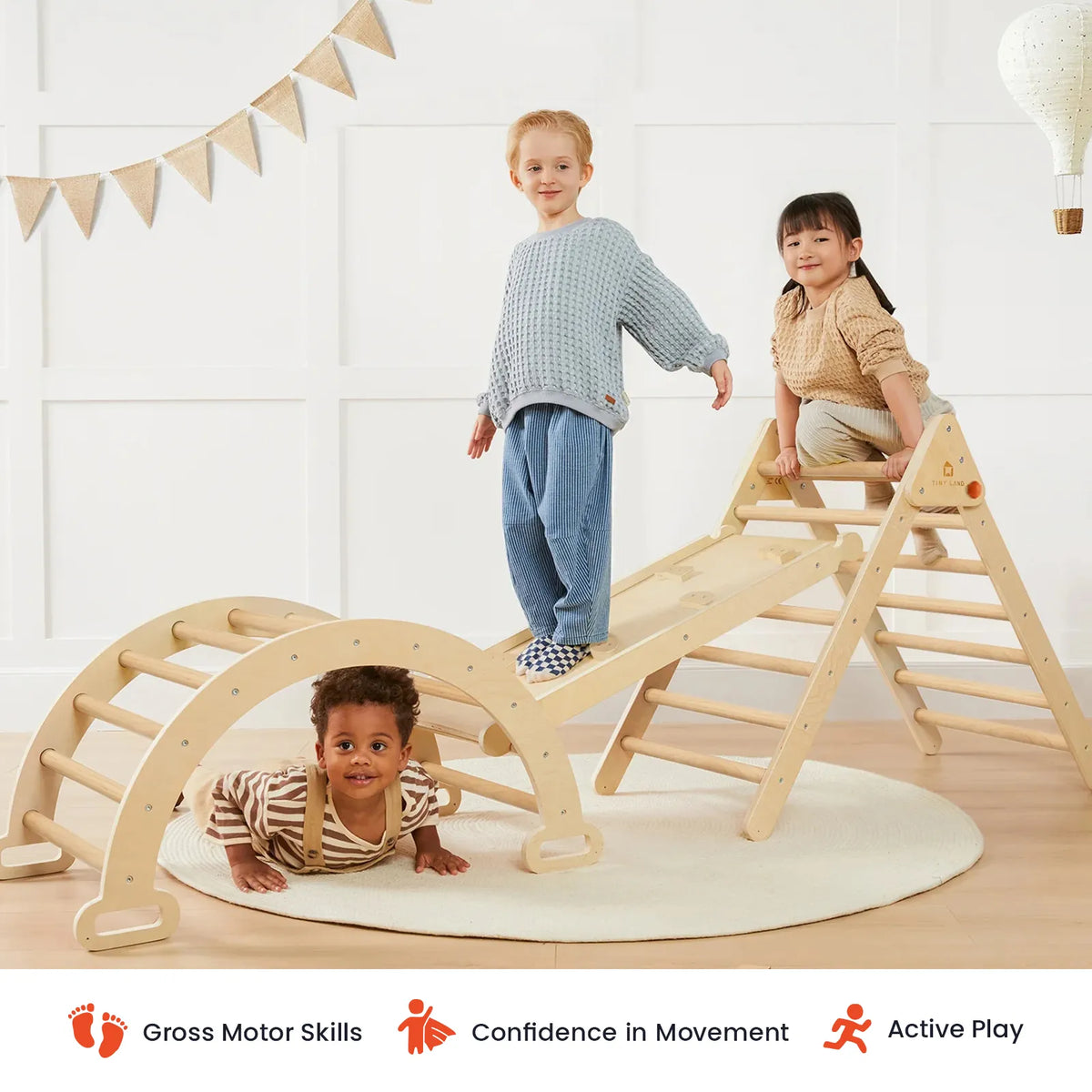 Three children enjoying indoor play on wooden climbing structures, including the Tiny Land® Pikler Triangle 7-in-1 Montessori Climbing Set; one child stands at the top of a ramp, another climbs, and a third crawls underneath. Text icons below emphasize gross motor skills, confidence in movement, and active play to support physical development—all with Tiny Land quality.|natural wood color