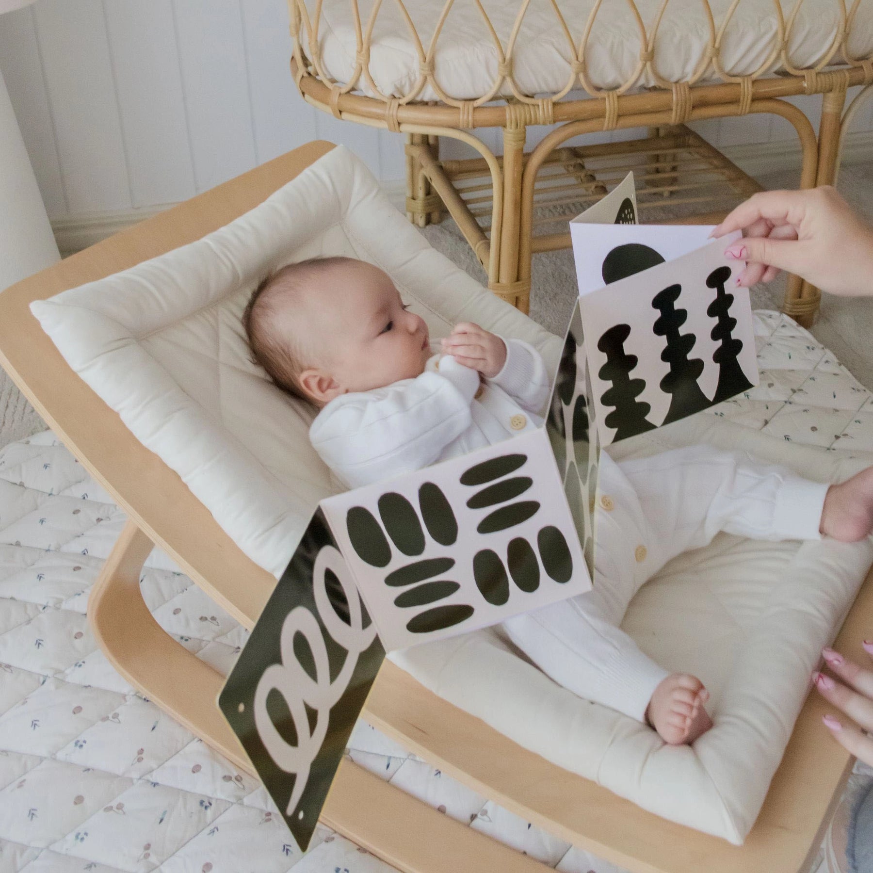 A newborn in a white onesie relaxes in a wooden rocker, looking at a black-and-white card from the Tiny Land® Montessori Toys for Newborns (0–3 Months) by Tiny Land Official Store, held by an adult nearby.