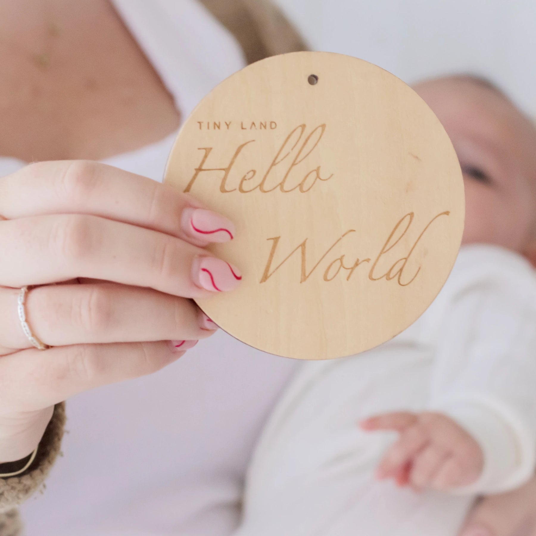 A person with pink and red wavy nails holds a round wooden sign reading “Tiny Land Hello World” in front of a newborn in white, featuring the Tiny Land® Montessori Toys for Newborns (0–3 Months) from Tiny Land Official Store.