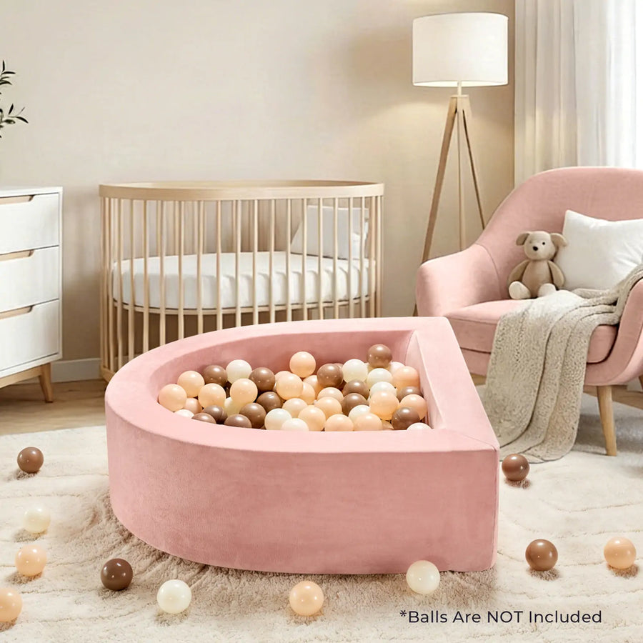 A modern nursery featuring the Tiny Land® PlayNest Soft Foam Ball Pit by Tiny Land, designed for sensory play and filled with beige, brown, and peach balls. The space also includes a wooden crib, a pink armchair with a teddy bear, a dresser, climbing blocks for playful exploration, a standing lamp, and scattered balls on the floor.|Pink