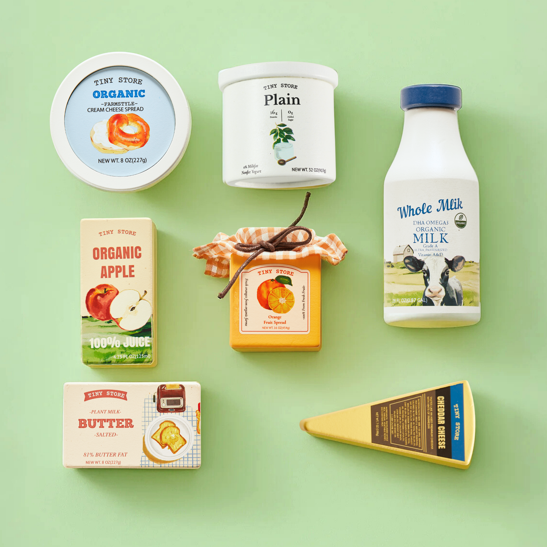 The Tiny Land® Organic Dairy Pack by Tiny Land INC features assorted dairy and apple-themed pretend food toys—milk, yogurt, juice, butter, cheese, and jam—arranged on a light green background with “Tiny Store” labels and colorful illustrations.