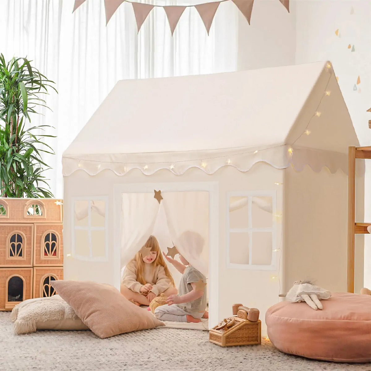 Two young children play inside the Tiny Land® Playhouse Tent – a natural cream indoor tent by Tiny Land – decorated with string lights, surrounded by soft pillows, a plush ottoman, wooden toys, plants, and bunting in a bright room.