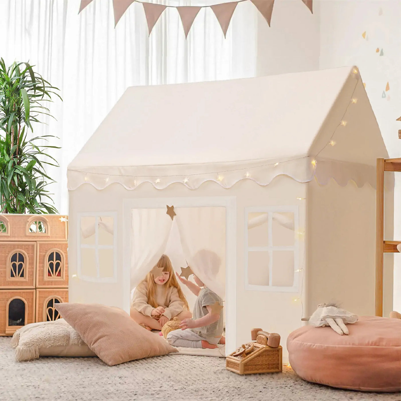 Two children enjoy the Tiny Land Bright Play & Art Set, featuring a colorful easel and a cream playhouse with lights, surrounded by pillows and toys in a bright, plant-filled room—perfect for imaginative indoor or outdoor fun.