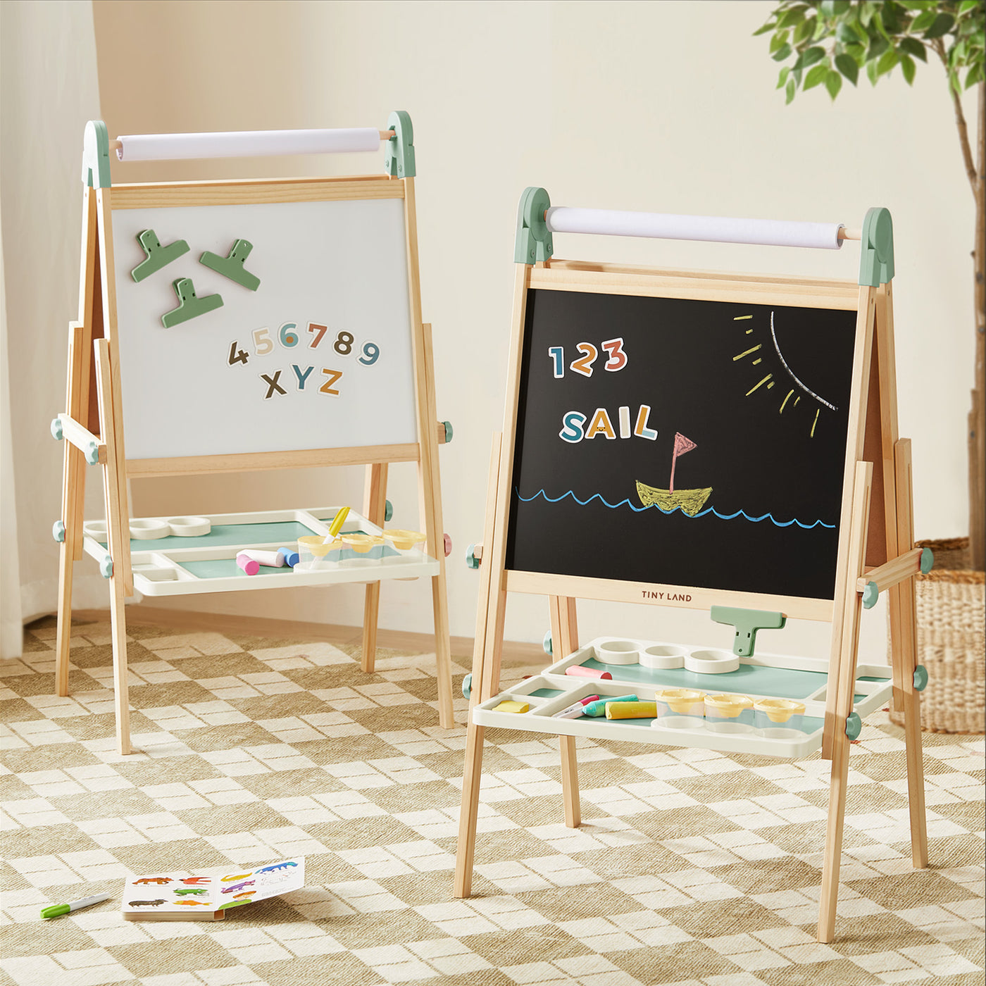Two Tiny Land® Magnitales Easel for Kids—one showing magnetic numbers and letters, the other with a chalk sailboat and "123 SAIL"—stand on a checkered rug. Art supplies rest on their trays, while a plant brightens the background.