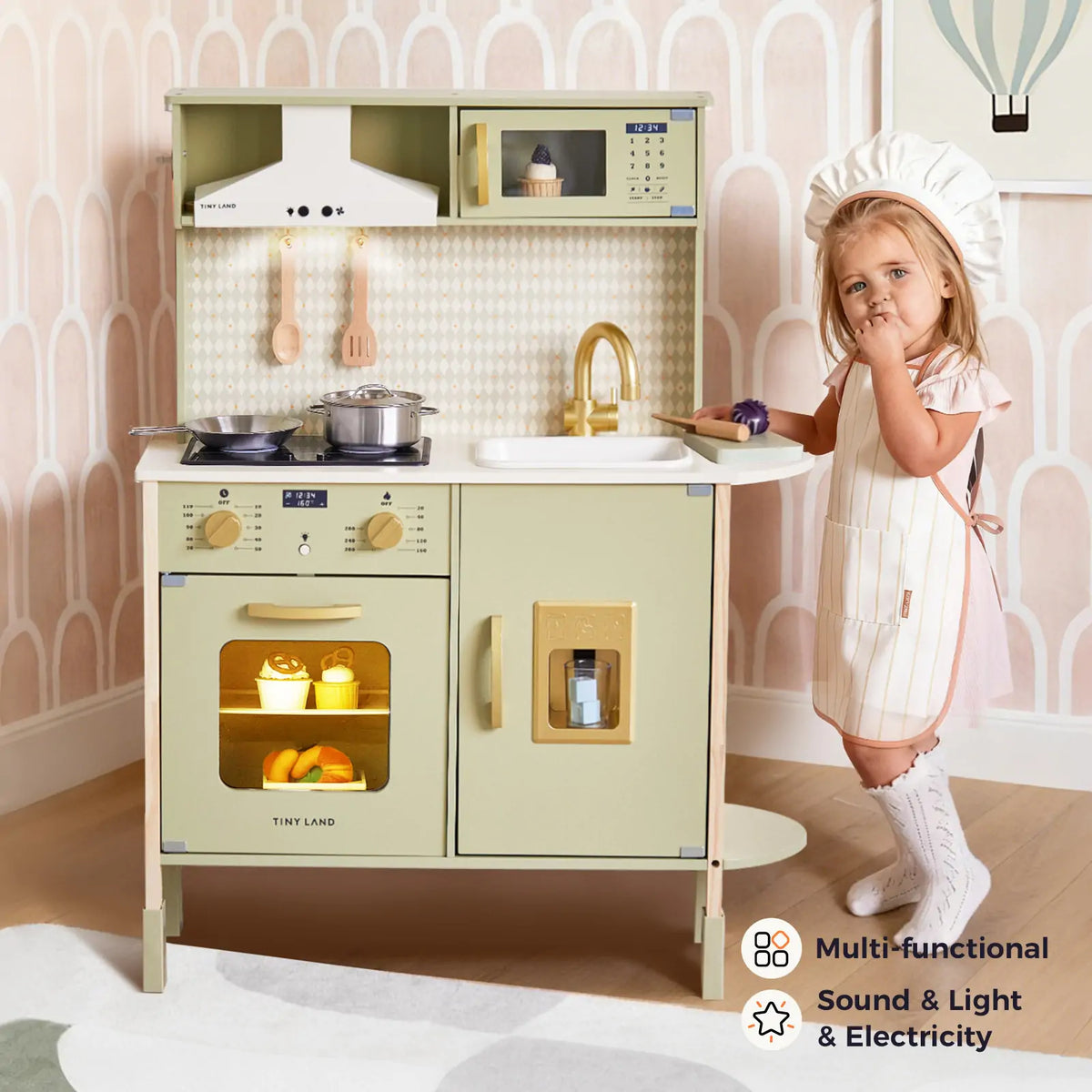 A girl in chef hat and apron stands by the Tiny Land® PopJoy Play Kitchen - Green, featuring a lit oven, sink, stove, play food, and multi-functional sound, light, and electricity features for realistic pretend play.