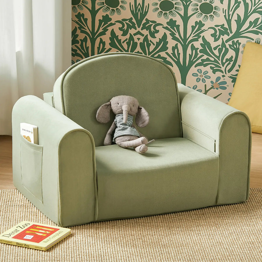 A soft green Tiny Land® GentleSnug Kid Chair with high-density foam and a handy side pocket holds a gray stuffed elephant toy. Children’s books are scattered on the wooden floor nearby, while green floral wallpaper and a partial yellow cushion provide a cozy backdrop.|Sage