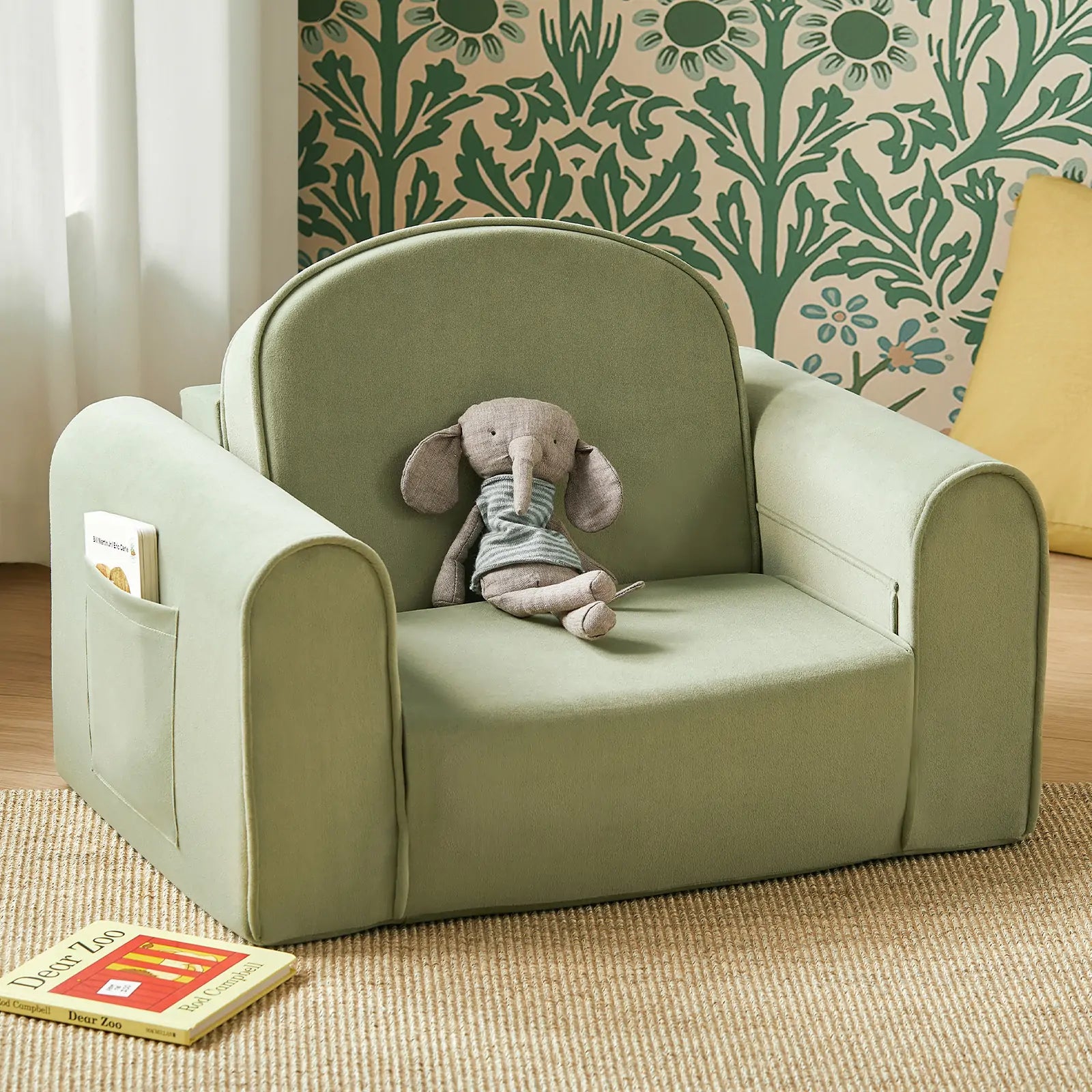 The Tiny Land® GentleSnug Kid Chair in Sage, by Tiny Land, features side pockets and a soft gray elephant toy in the center. A book lies on the floor, while green floral wallpaper adds to the cozy, inviting atmosphere.