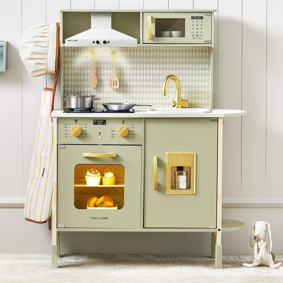 The Tiny Land® PopJoy Play Kitchen - Green by Tiny Land features a pastel green wooden design with gold accents, stovetop, oven with window, microwave, sink, hanging utensils, and play food. A chef’s hat and stuffed rabbit rest on the light floor nearby.