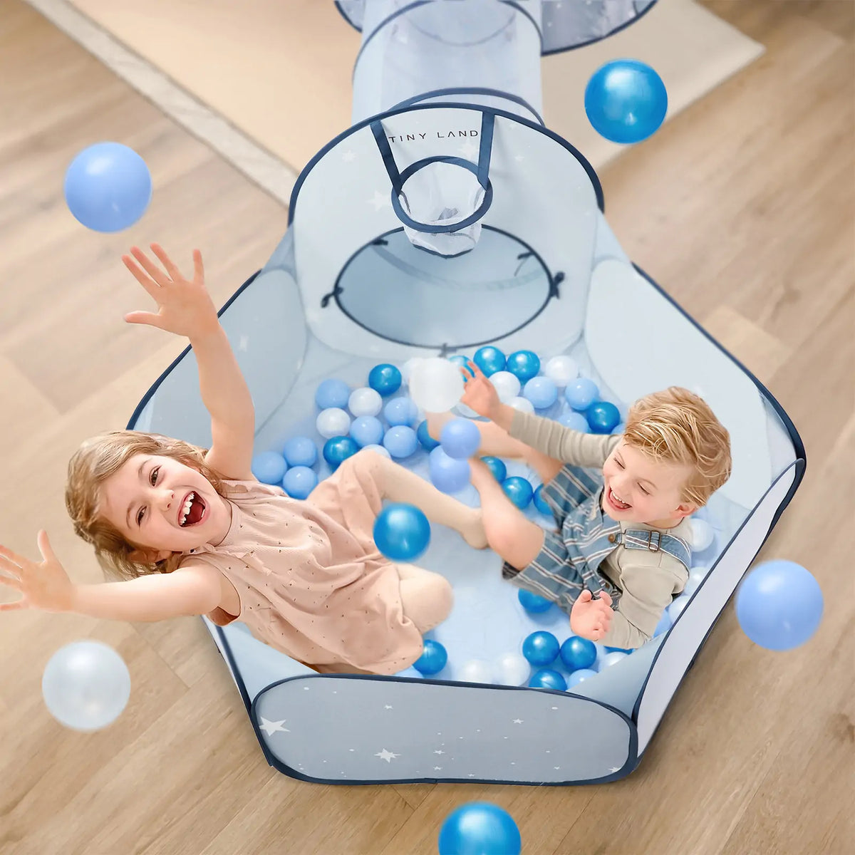 Two young children play joyfully in a small ball pit filled with Tiny Land® Blue Ocean Balls (200 pcs) by Tiny Land, with one child tossing a ball. The light blue play area features a mesh tunnel overhead.|blue