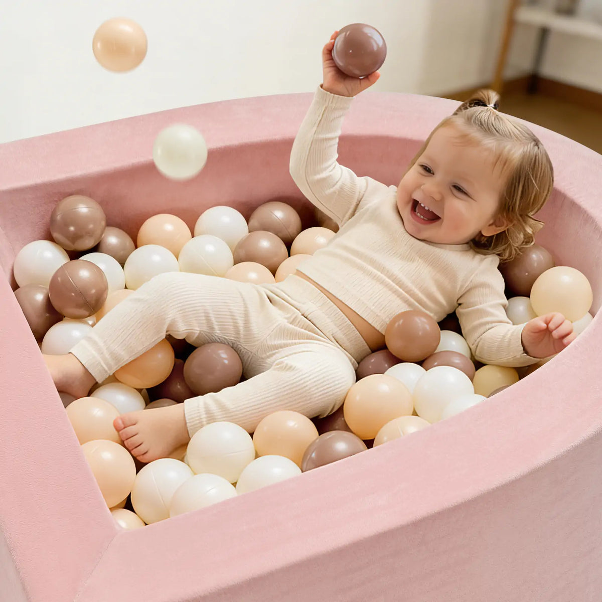 A smiling toddler enjoys sensory play in the Tiny Land® Pink PlayNest Ball Pit by Tiny Land INC, holding a ball and looking happy in cozy cream-colored clothes.|pink