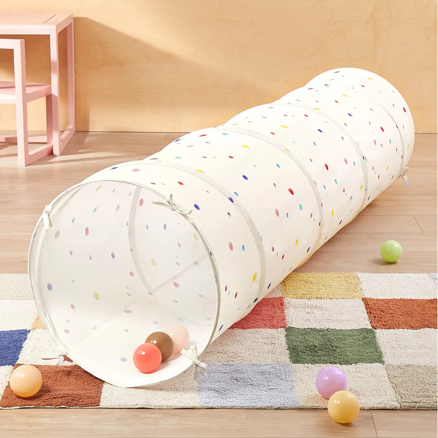 The Tiny Land® Adventure Play Tunnel - Colorful Polka Dots by Tiny Land is set on a multicolored checkered rug with small colorful balls, while a pink stool stands near a light wood wall in the background.