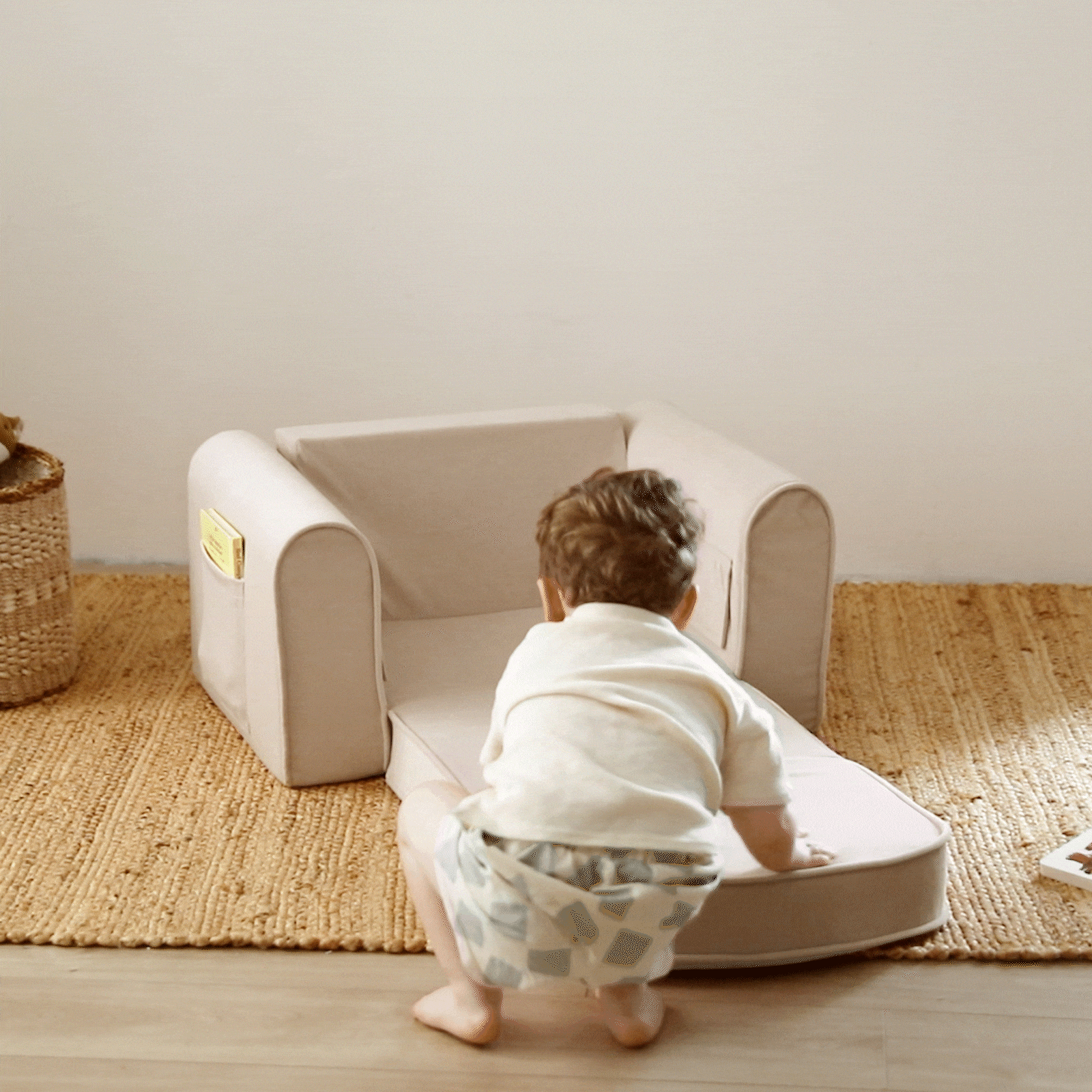 A toddler in a white shirt and patterned shorts kneels on a beige rug, unfolding the Tiny Land® GentleSnug Kid Chair - Beige White by Tiny Land into a small lounger in a cozy room.