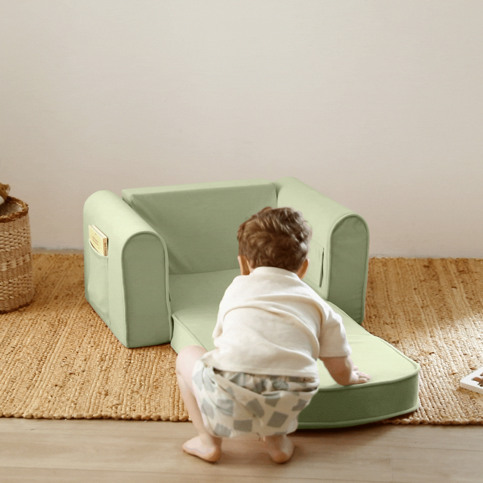A toddler in a white shirt and patterned shorts squats by the Tiny Land® GentleSnug Kid Chair - Sage, a foldable kids sofa with footrest from Tiny Land, placed on a woven rug in a cozy room.