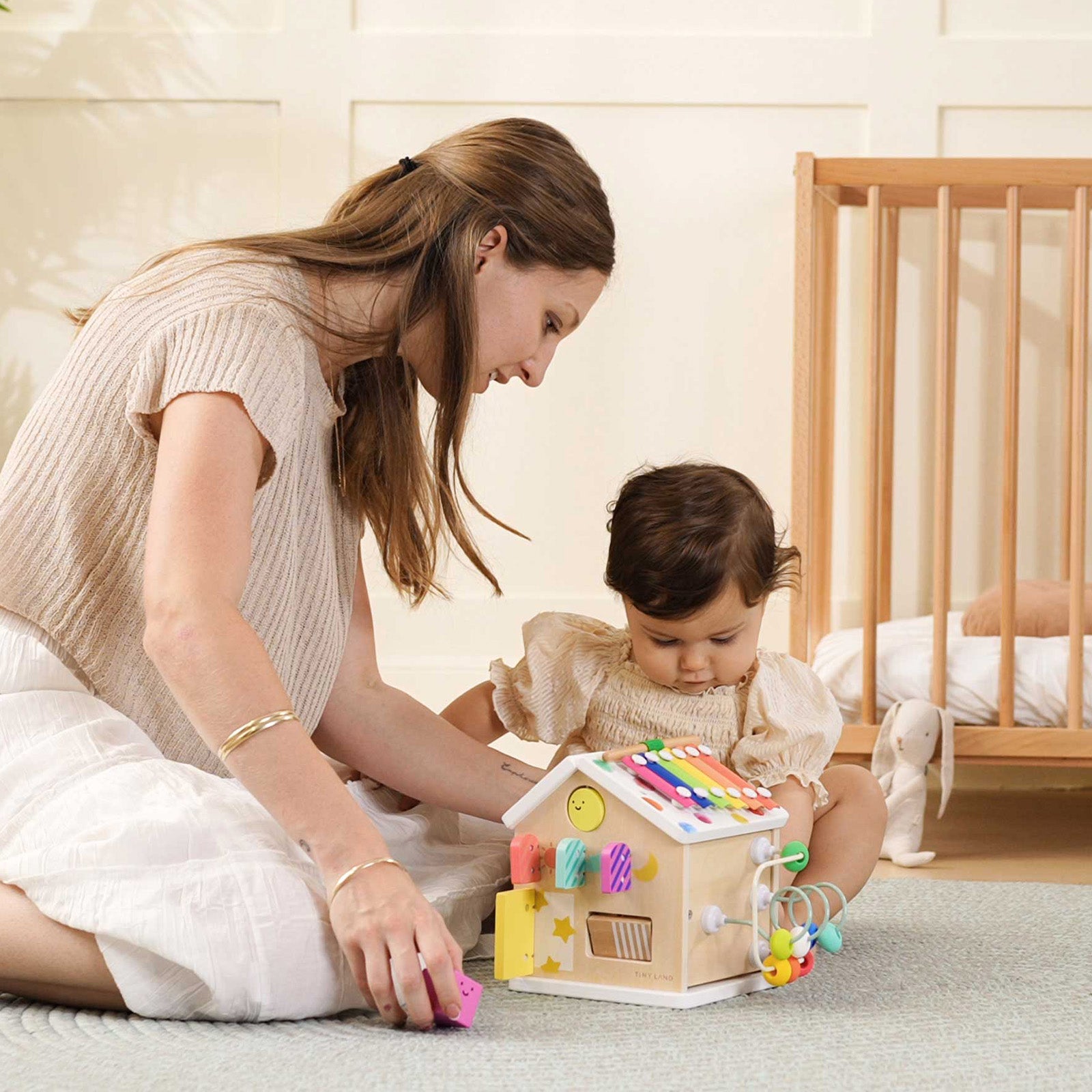 A woman and a young child sit on the floor playing with the Tiny Land® Cottage Activity Cube - Rainbow by Tiny Land in a softly lit, cozy nursery with a crib in the background.