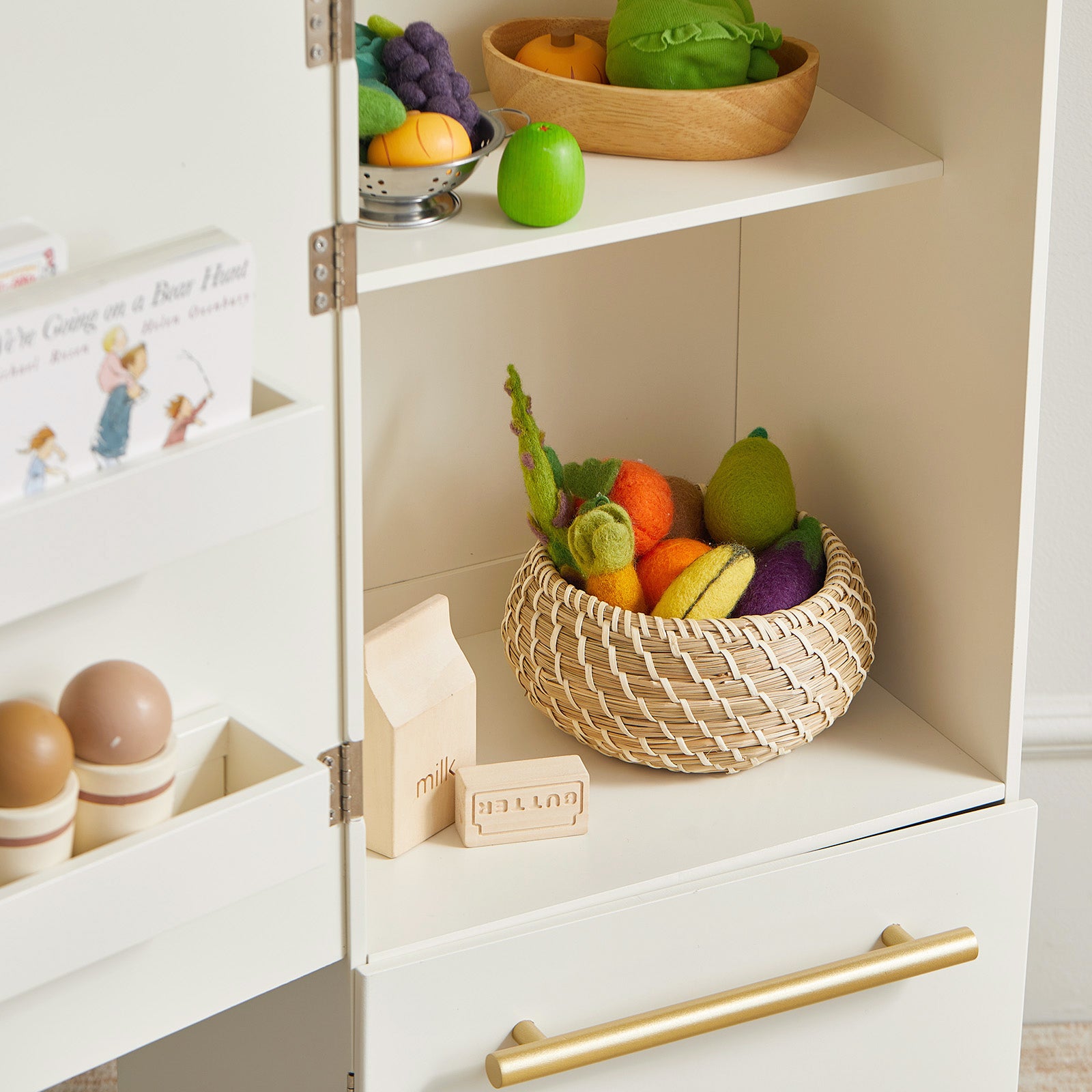 The Tiny Land® Play Kitchen Accessories – Fridge is a white wooden cabinet with shelves that holds toy food, including plush veggies, a green apple, wooden butter block, eggs, and a storybook—ideal for any play kitchen.
