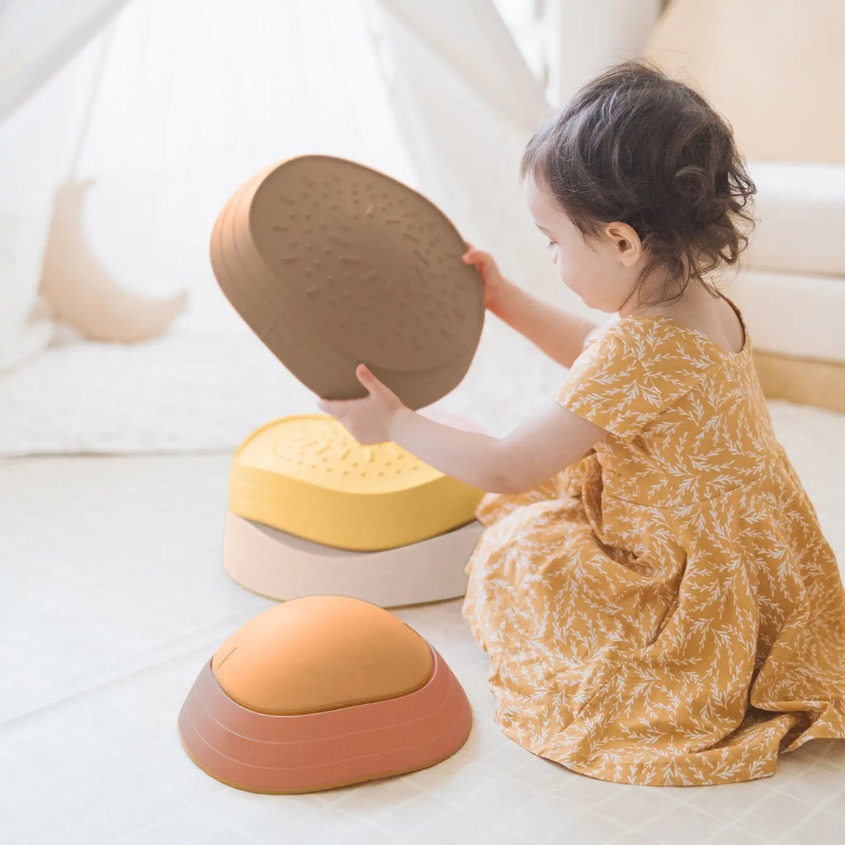 A young child in a yellow patterned dress sits on the floor, stacking Tiny Land® Stepping Stones - Sunset Hues, developing balance and coordination in a bright, cozy room.|Sunset Hues