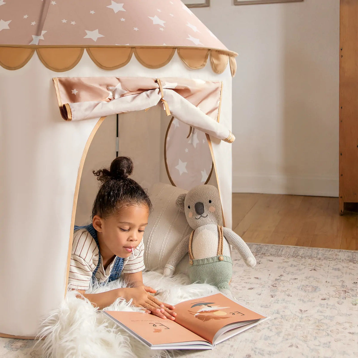 A young child reads a picture book inside the Tiny Land® Kids Tent from Target, lying on the floor with a plush bear on a cozy rug as soft natural light brightens the star-patterned indoor tent for toddlers.|brown