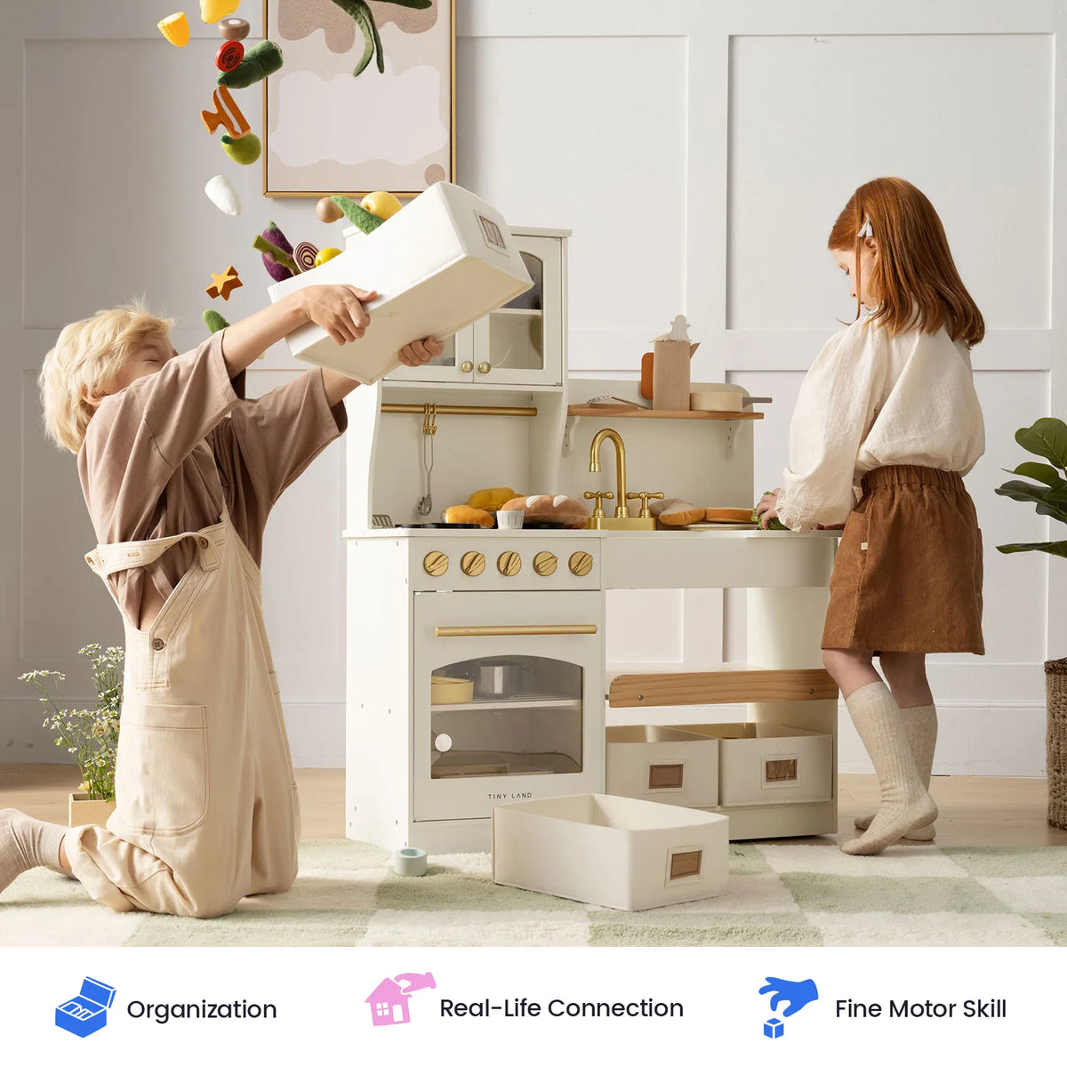 Two children play with the Tiny Land® Wooden Play Kitchen with Storage for Creative Toddlers; one pours toy vegetables from a storage bin while the other stands nearby. Pretend cookware, bins, and play food are scattered on the floor. Icons below highlight organization, real-life connection, and fine motor skill development.|cream