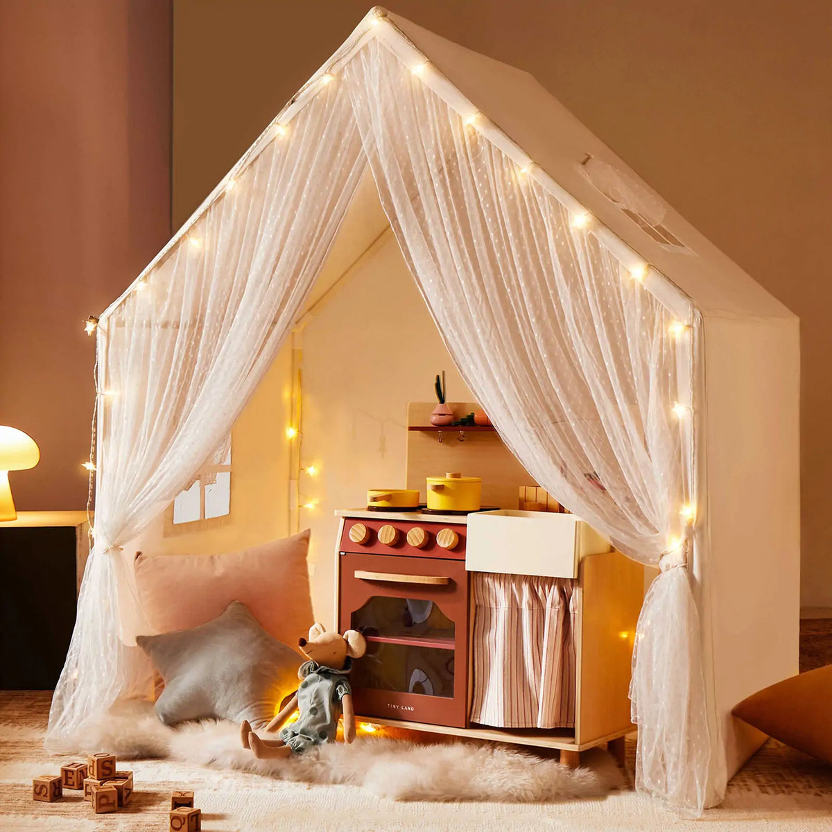 The Tiny Land® Large Space Play House with Star Lights by Tiny Land is a cozy kids’ playhouse featuring string lights, sheer curtains, a toy kitchen, plush cushions, soft rug, wooden blocks, and a stuffed mouse for inviting play.