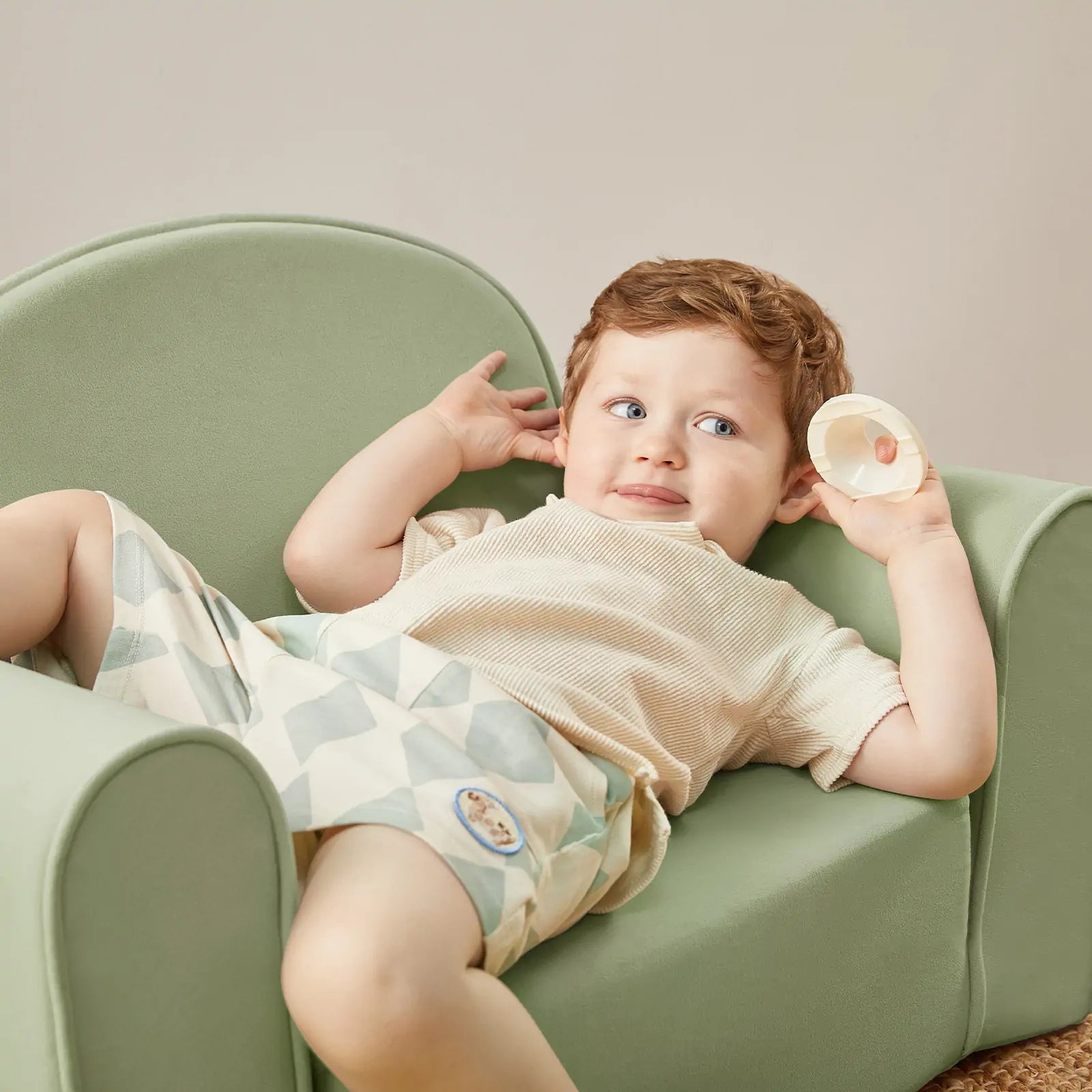 A young child lounges on the Tiny Land® GentleSnug Kid Chair in Sage by Tiny Land, wearing a beige shirt and patterned shorts, holding a small white cup and looking playfully to the side.