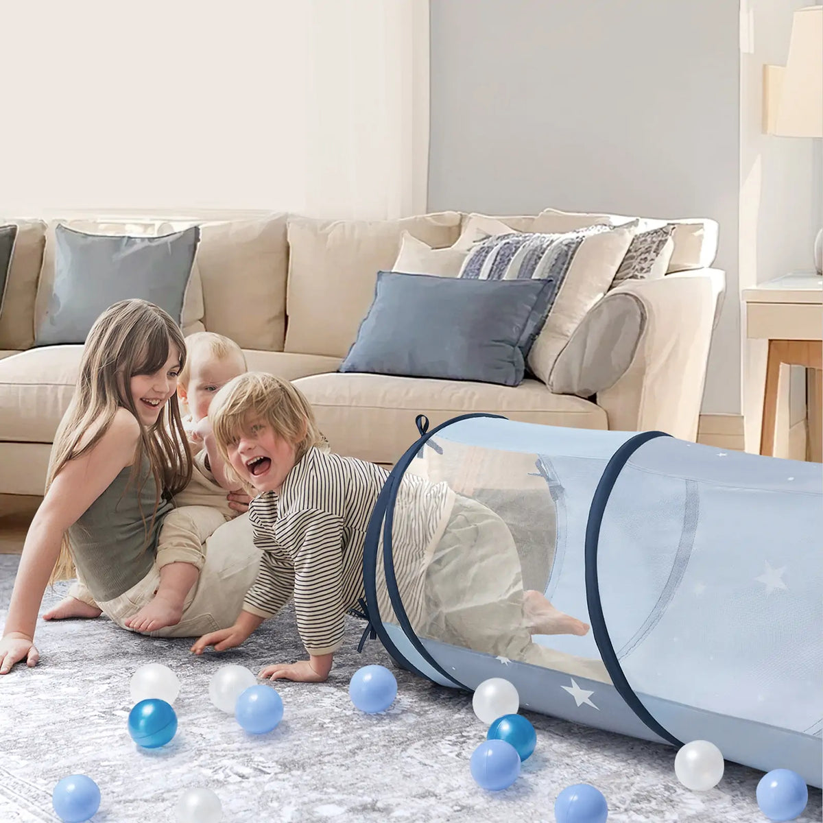Two young children play on a living room rug with the Tiny Land® Adventure Play Tunnel - Blue by Tiny Land and scattered blue and white plastic balls, while a beige couch with blue pillows sits in the background.