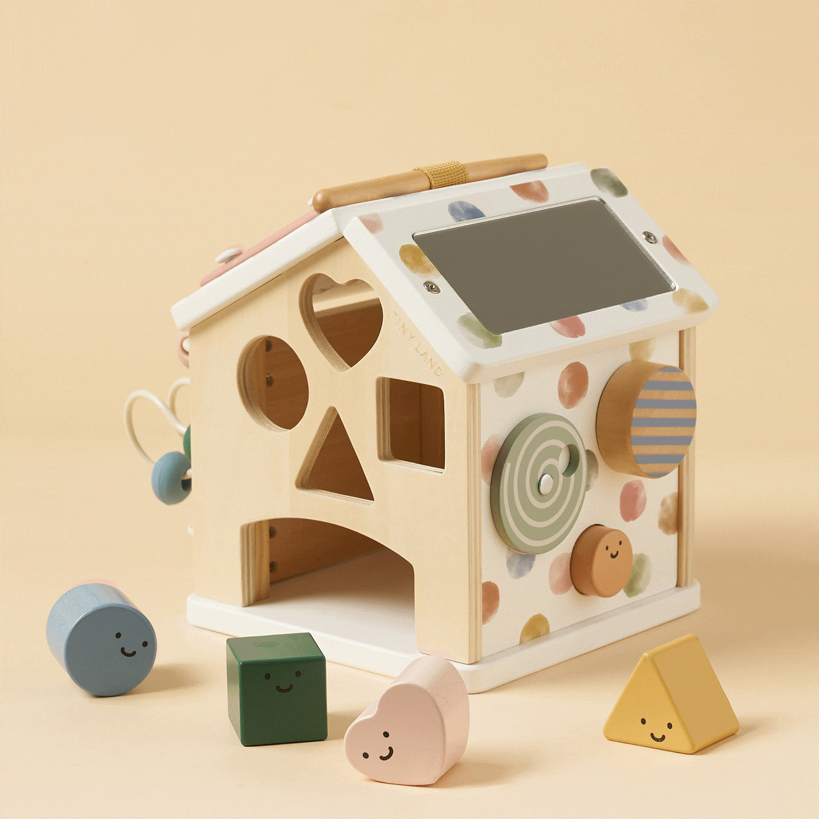 The Tiny Land® Cottage Activity Cube - Morandi by Tiny Land is a wooden Montessori toy house with pastel heart, square, circle, and triangle shapes featuring smiley faces, matching cut-out holes, and a small chalkboard roof.
