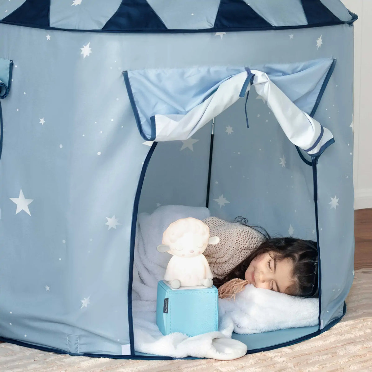 A young child sleeps inside the Tiny Land® Kids Tent – Starry Blue Indoor Tent for Toddlers, resting on a white pillow and blanket. A soft toy and blue music box are placed near the entrance of this cozy tent by Tiny Land.