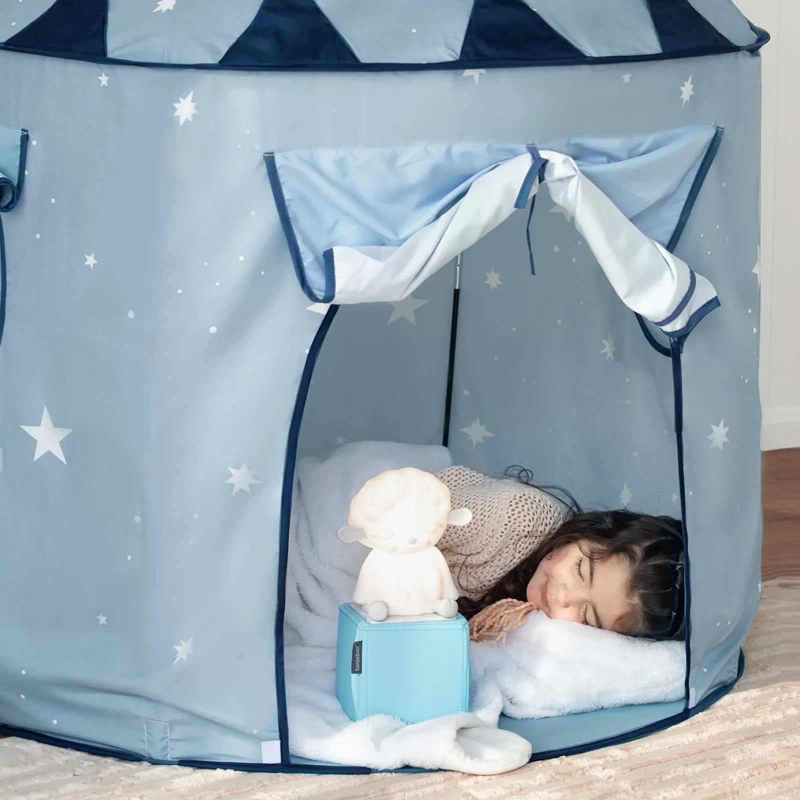 A young child sleeps inside the Tiny Land® Kids Tent – Starry Blue Indoor Tent for Toddlers, resting on a white pillow and blanket. A soft toy and blue music box are placed near the entrance of this cozy tent by Tiny Land.