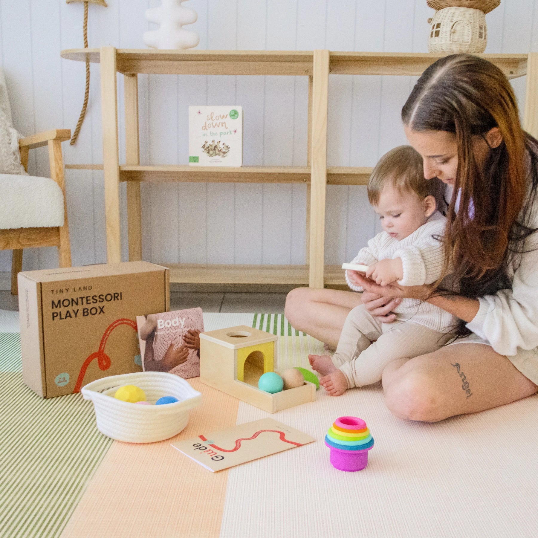 A woman and a baby sit on a play mat with Tiny Land® Montessori Toys for Newborns (7–9 Months), including stacking cups, a sensory ball, an Object Permanence Box, and books. The open Tiny Land box is beside them with shelves in the background.