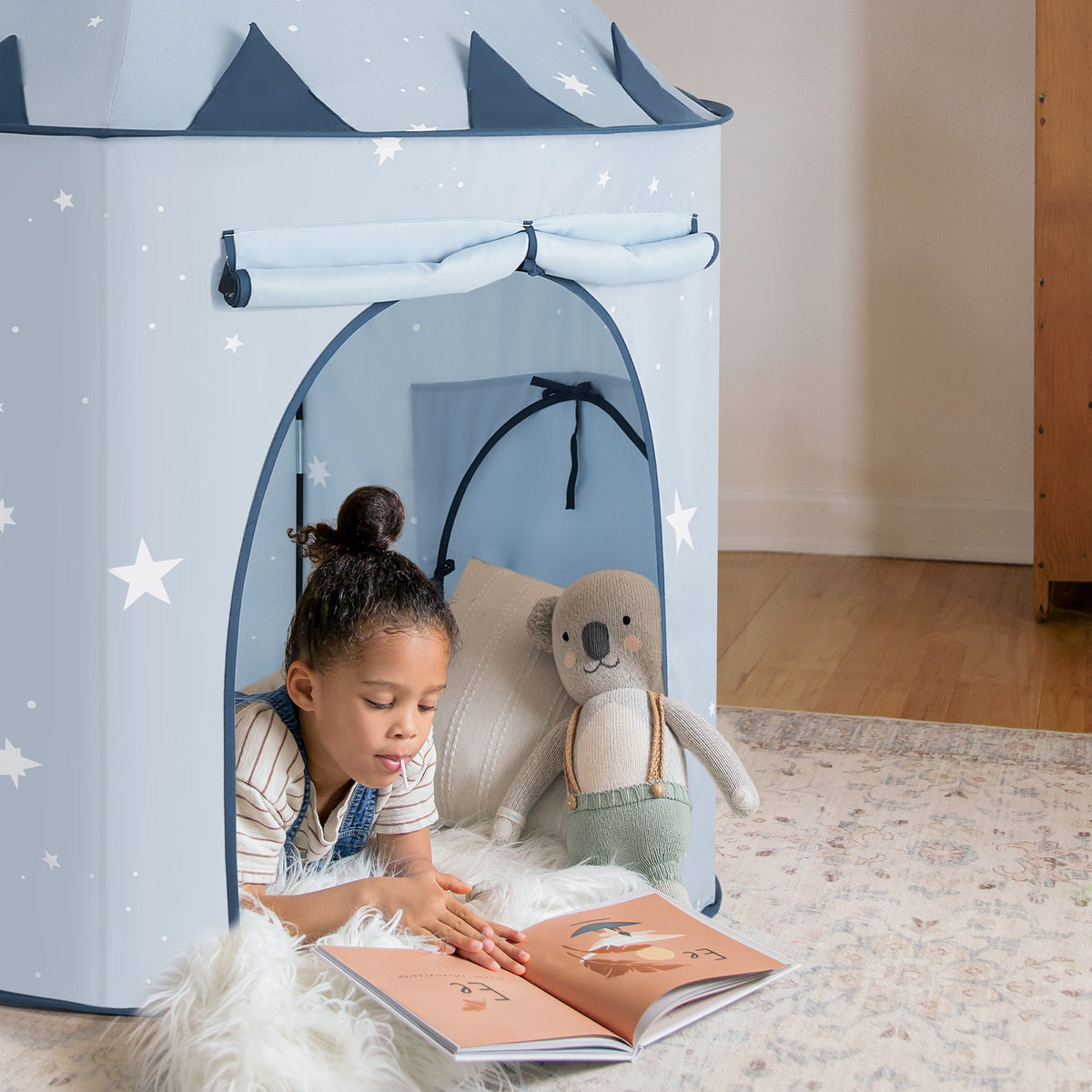A young girl enjoys reading inside the Tiny Land® Adventure 3-in-1 Play Tunnel & Ocean Ball Set - Blue, with a stuffed koala beside her and a matching tunnel nearby, all on a soft, light-colored rug.|blue