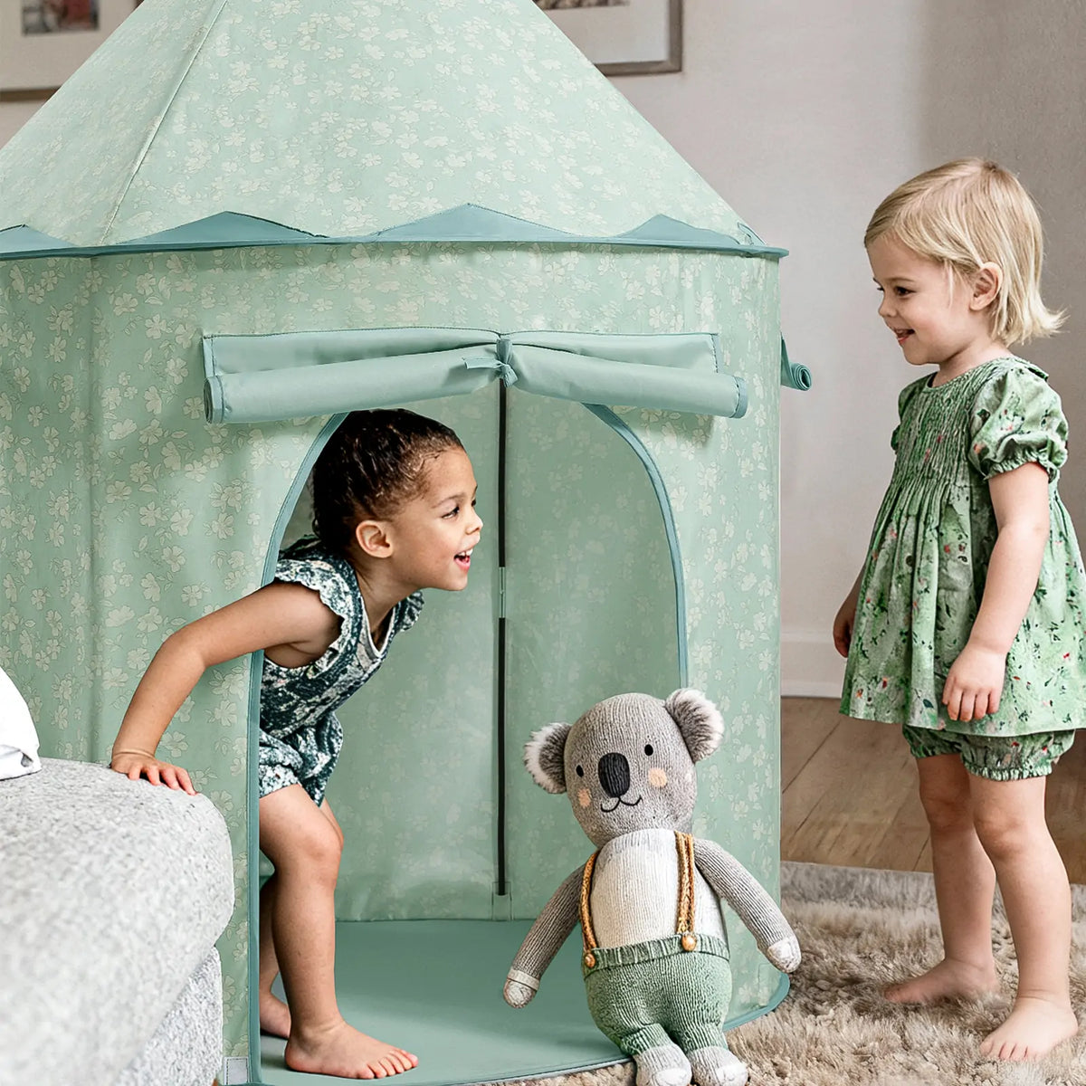 Two young children play happily near the Tiny Land® Tiny Sprout Castle Tent in pastel green. One child crouches at the tent entrance, smiling, while the other stands nearby. A plush koala toy with suspenders sits in front.|green