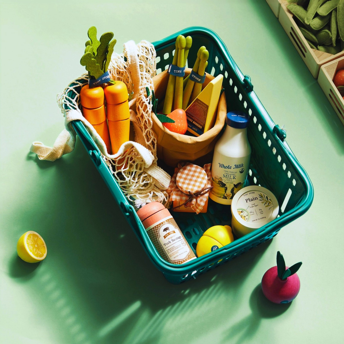 A green shopping basket holds the Tiny Land® Organic Play Food Set by Target, featuring plush toys and felt models of carrots, asparagus, milk, yogurt, a jar, lemon, and apple, all displayed on a light green surface.